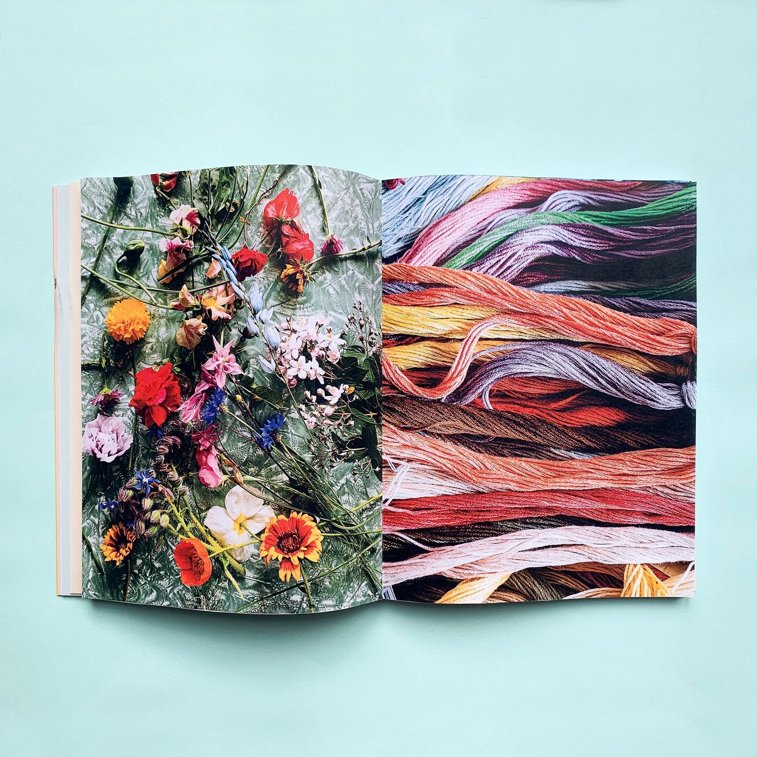 An open book or art print laid flat on a light aqua background, showing a floral photograph on the left page and a close-up of multicolored yarn or thread skeins on the right page.