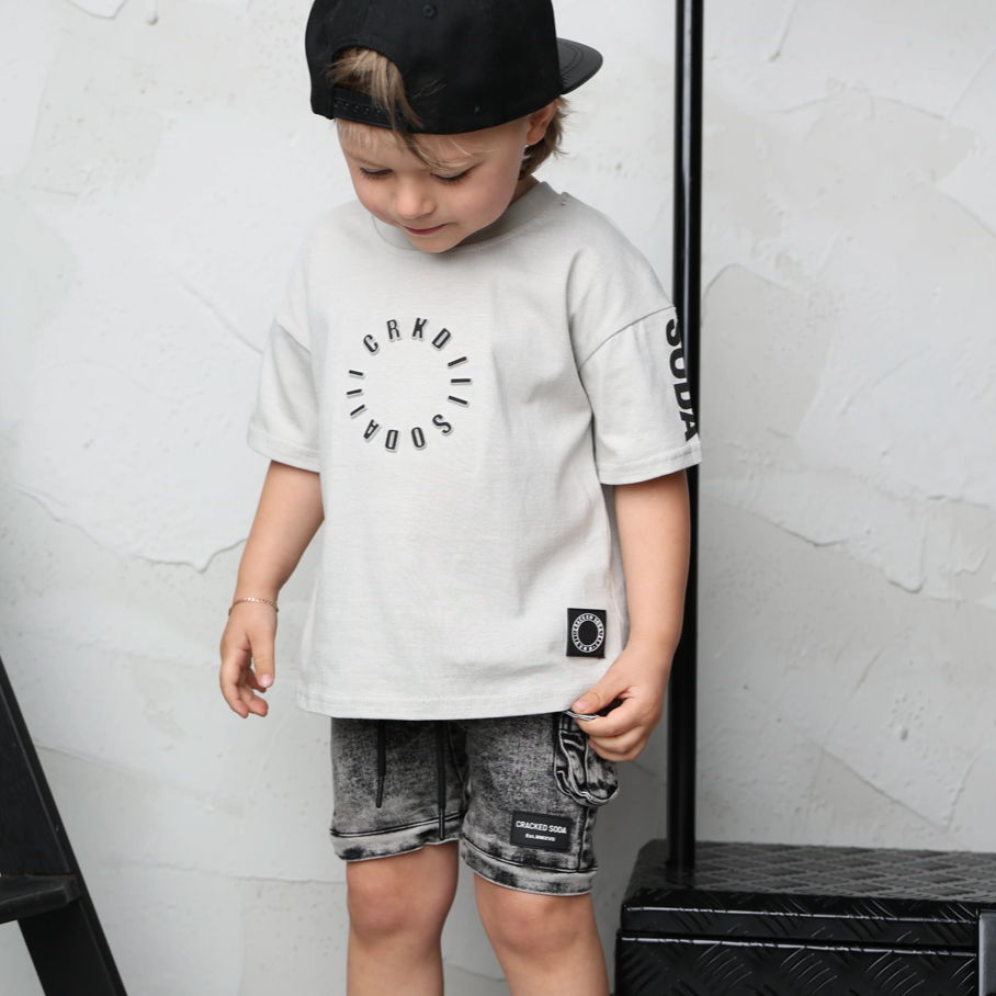Child wearing a white t-shirt with black graphics, black cap, and gray shorts standing indoors.
