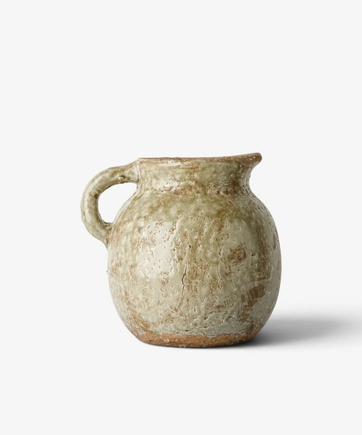 A rustic handmade ceramic pitcher with a rounded body, a small loop handle, and a short spout, featuring a textured glaze in muted beige, olive green, and brown tones, set against a light gray background.