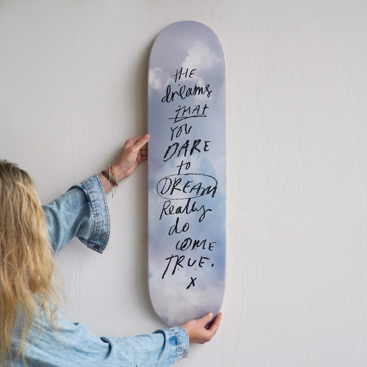 A person in a light blue denim jacket holds a vertical skateboard wall art piece against a white textured wall. The skateboard features a dreamy, cloudy sky design in soft lavender and blue-gray tones, with the handwritten quote 'THE dreams THAT YOU DARE to DREAM Really do COME TRUE.