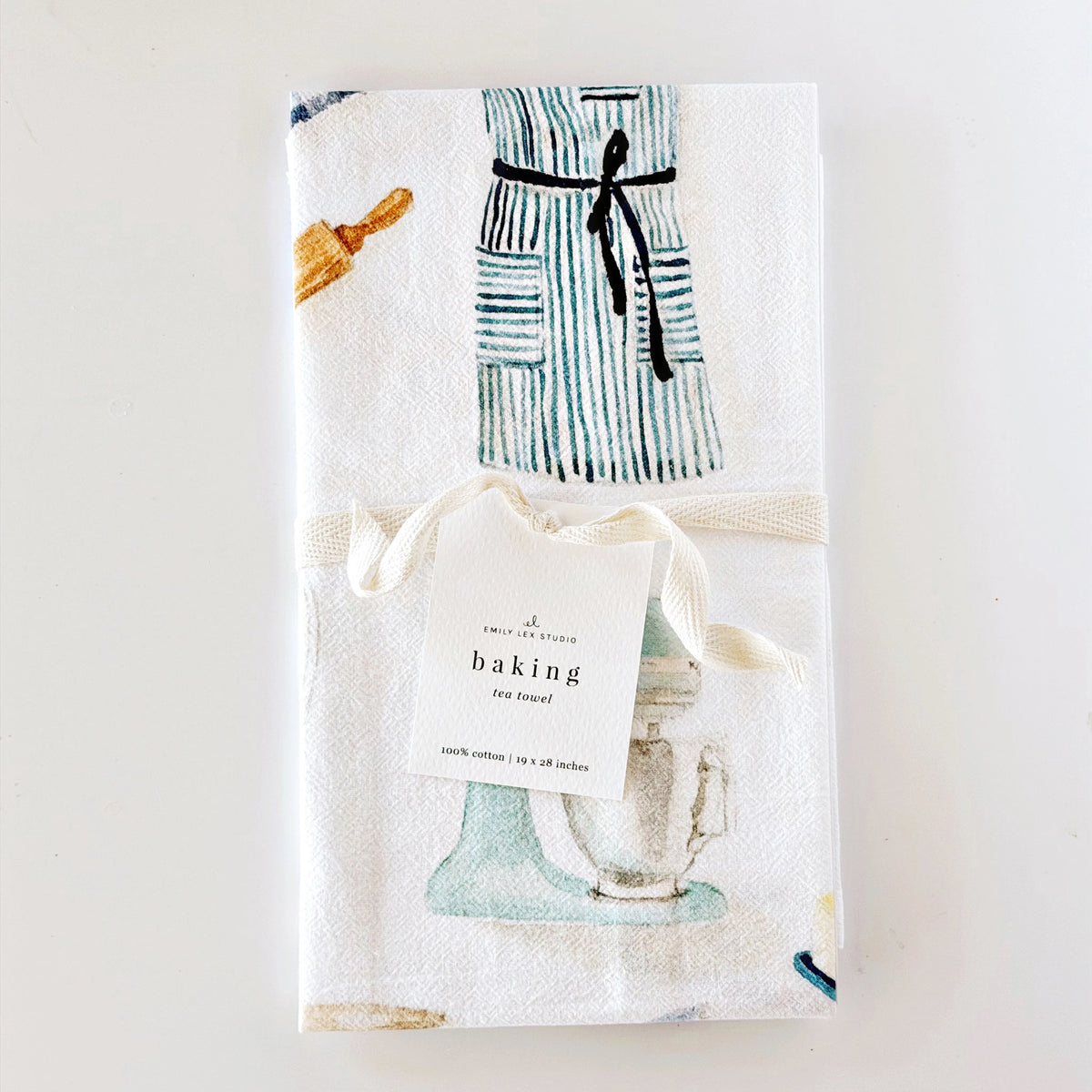 A folded white kitchen towel with a watercolor-style print of a striped apron, rolling pin, and stand mixer, tied with a cream ribbon and a paper tag that reads 'baking tea time!'.