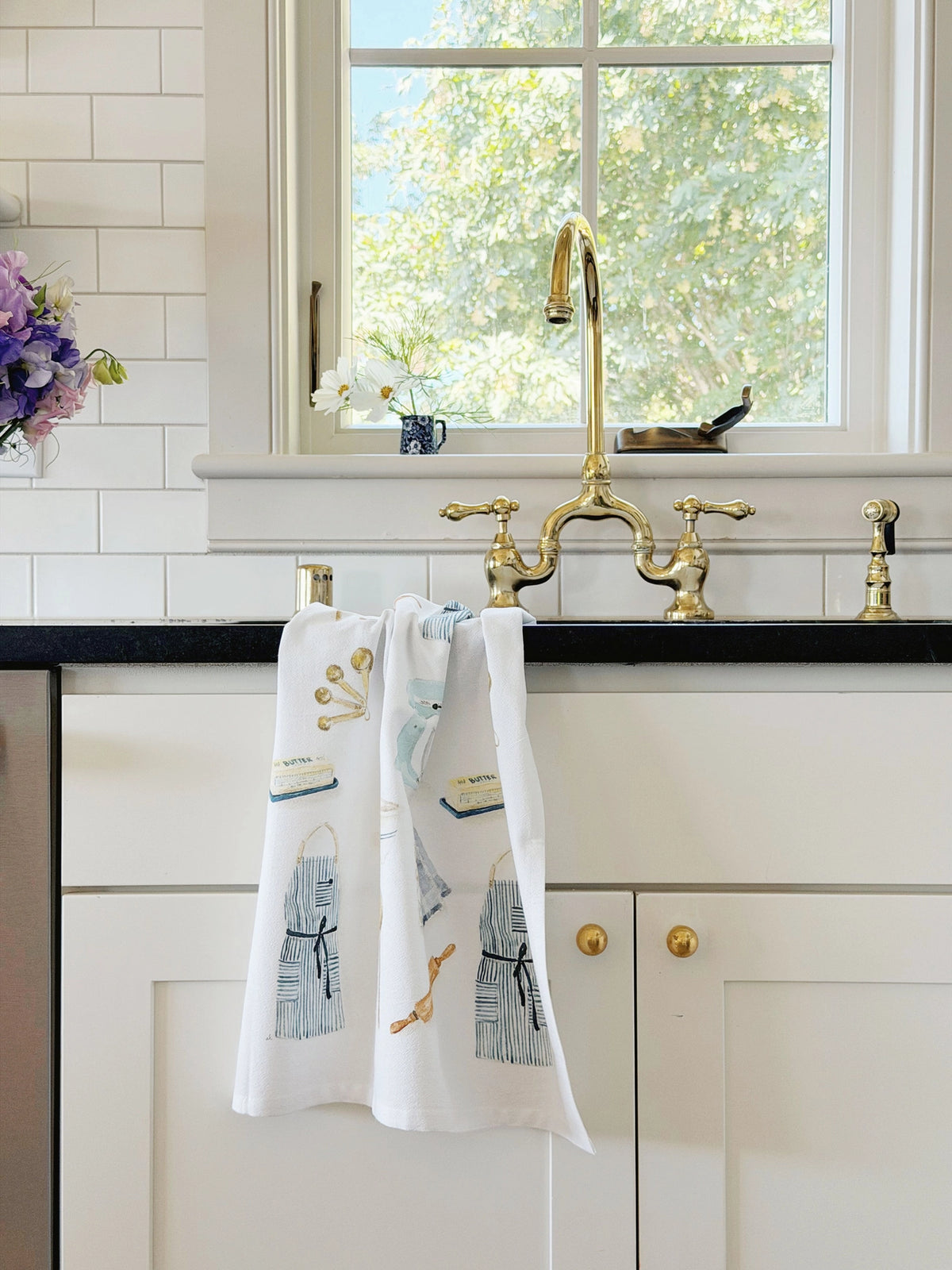 A white kitchen towel with a blue and gold nautical print hangs from a brass faucet in a bright kitchen with white subway tiles and a black countertop.