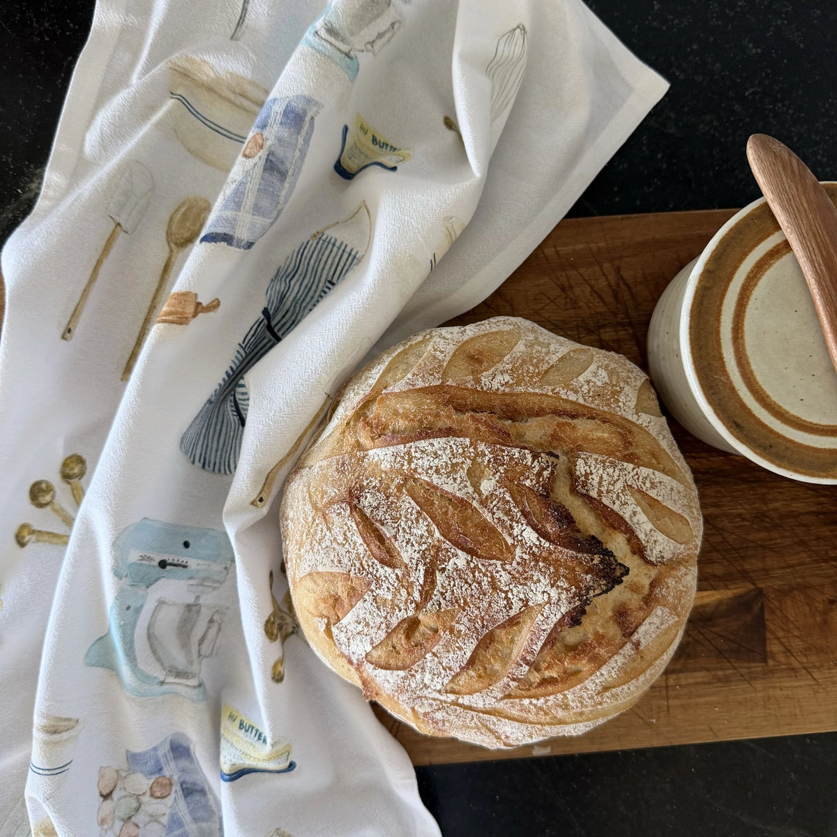 A round artisan bread loaf with flour-dusted scoring sits on a wooden cutting board next to a white tea towel printed with kitchen utensil illustrations, with a ceramic bowl and wooden spoon also visible.
