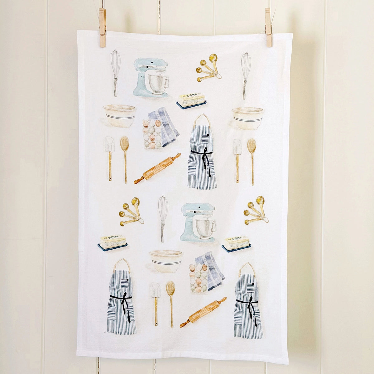 A white kitchen towel with a printed pattern of baking tools and ingredients, hanging from two wooden clips against a light beige paneled wall.