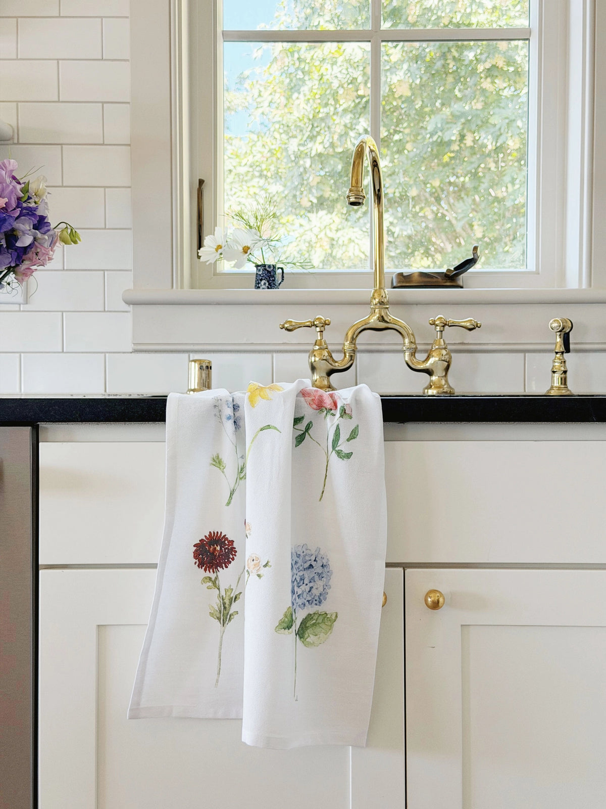 A white kitchen towel with a colorful floral print hangs from a brass faucet in a bright, modern kitchen with white subway tiles and a black countertop.