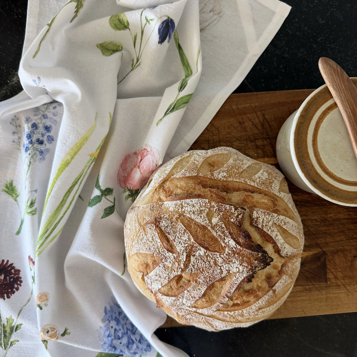 A round artisan bread loaf with a flour-dusted crust sits on a wooden board next to a white floral tea towel and a ceramic cup with a wooden spoon.