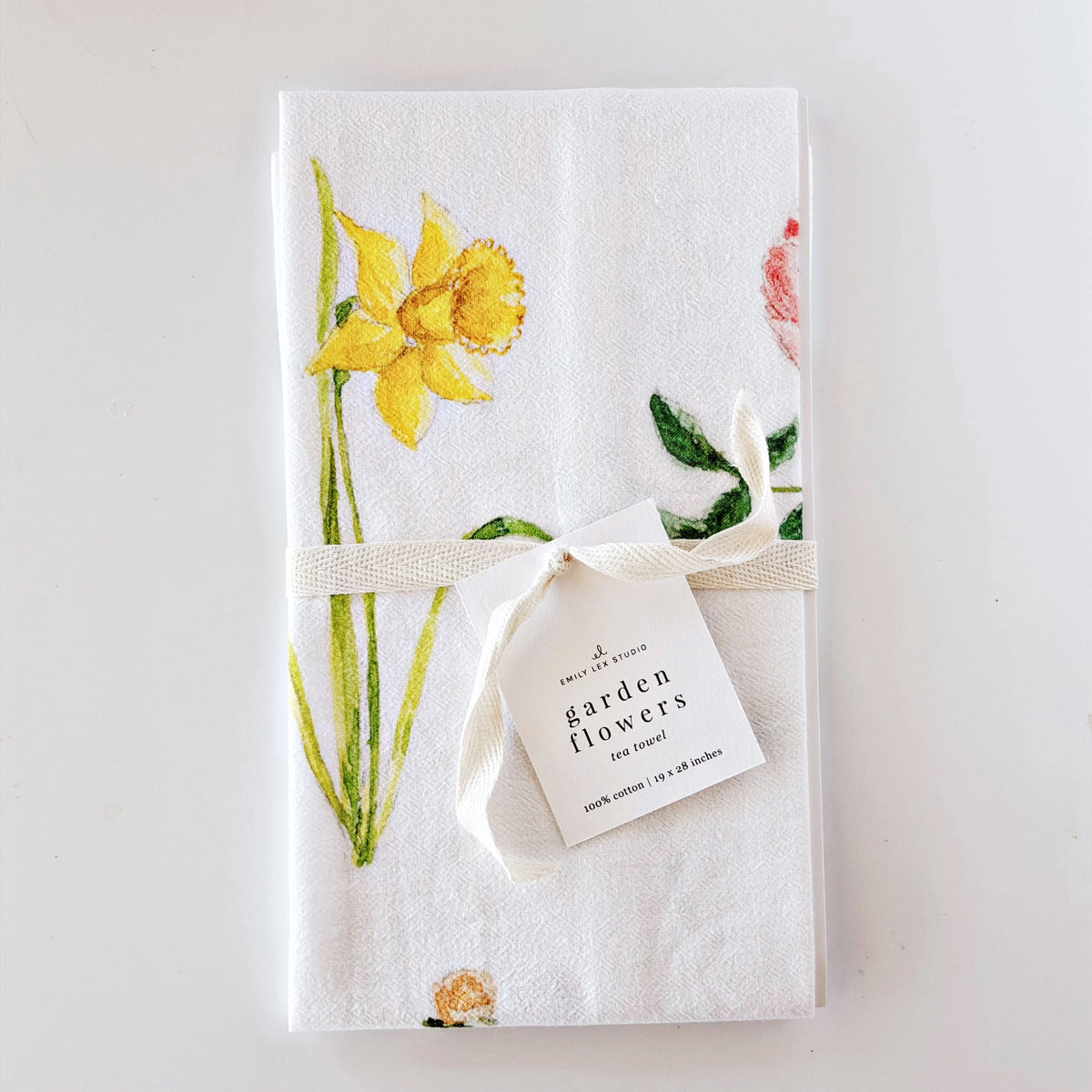 A white cotton tea towel featuring a watercolor print of yellow daffodils and green leaves, tied with a cream ribbon and a paper tag that reads 'garden flowers tea towel'.
