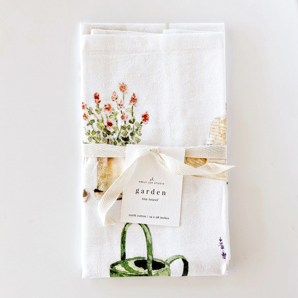 A folded white cotton tea towel with a garden-themed print, tied with a cream ribbon and a paper tag that reads 'garden tea towel'. The print features red flowers in a pot, a green watering can, and a seashell.