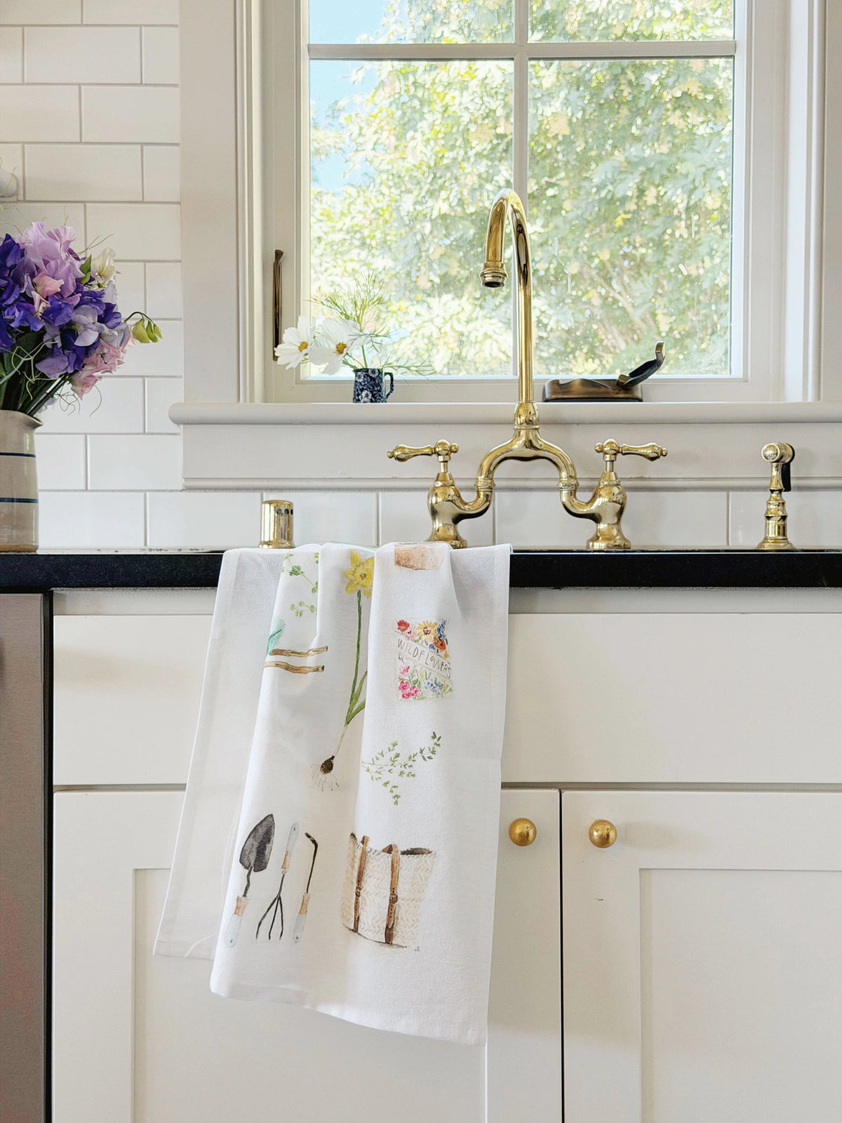 A white kitchen towel with colorful gardening illustrations hangs from a brass faucet in a bright kitchen with white subway tiles, a black countertop, and a window.