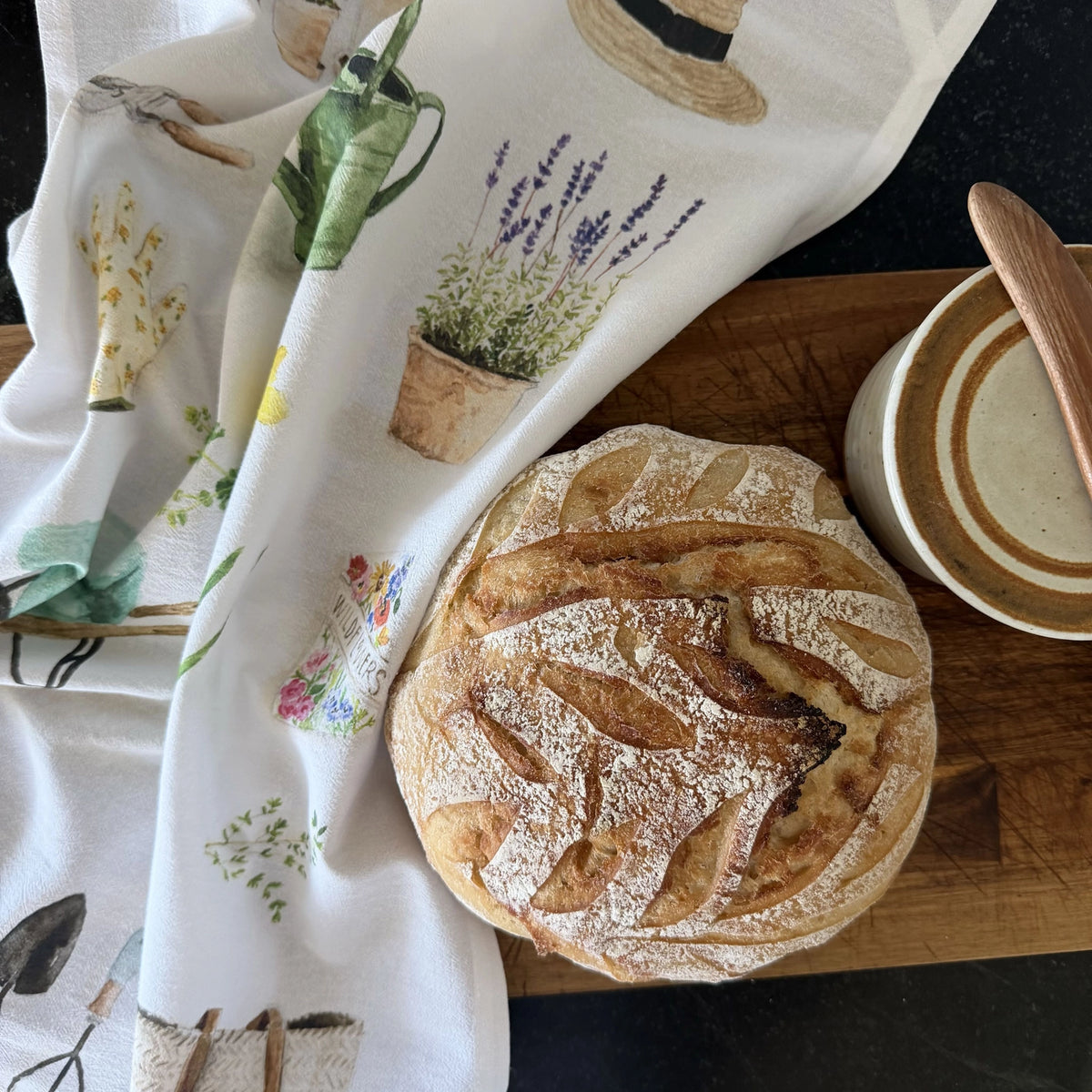 A round artisan bread loaf sits on a wooden board next to a white tea towel printed with botanical illustrations, with a ceramic bowl and wooden spoon also visible.