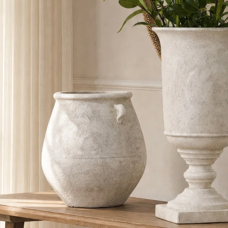 A textured light gray stone-look urn-shaped planter with a small side loop, displayed on a wooden table next to a larger matching pedestal planter with green foliage.
