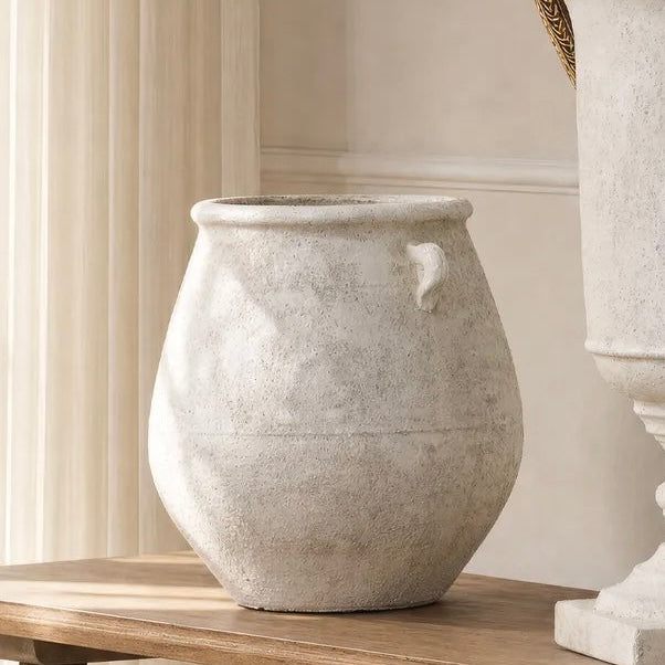 A textured light gray stone-look urn-shaped planter with a small side loop, displayed on a wooden table next to a larger matching pedestal planter with green foliage.