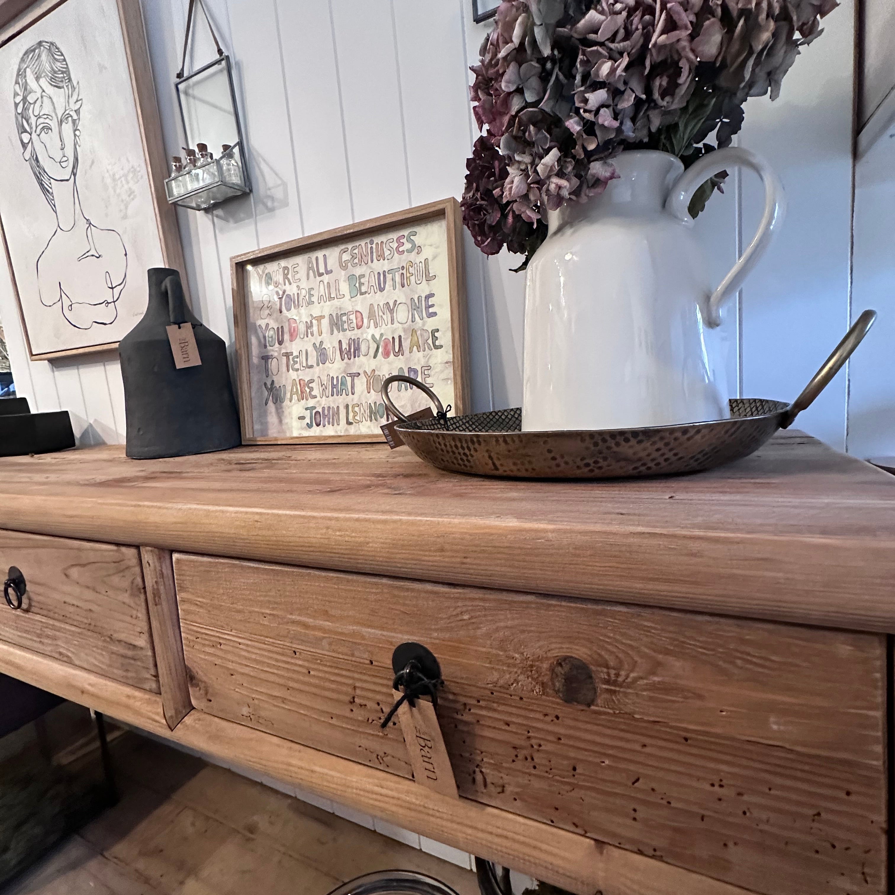 A rustic wooden console table with two drawers, styled with a white pitcher of dried purple flowers, a metal tray, and framed wall art in a home decor setting.