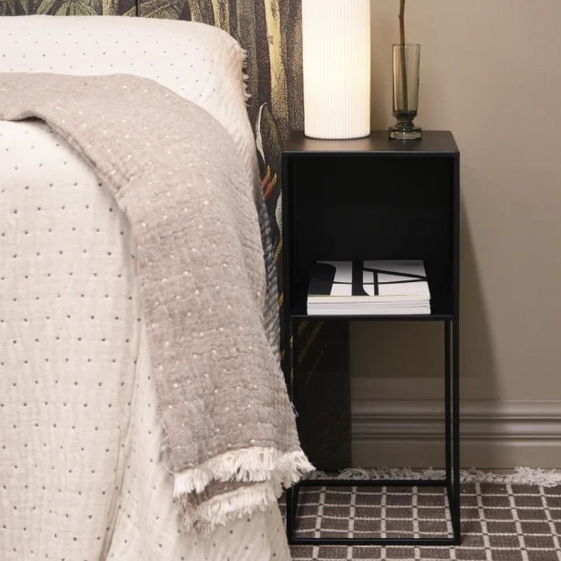 A black metal nightstand with an open shelf styled in a bedroom next to a bed with a textured cream quilt and a beige throw blanket. A white ribbed lamp and a dark glass vase sit on top of the nightstand, which also holds a stack of books.