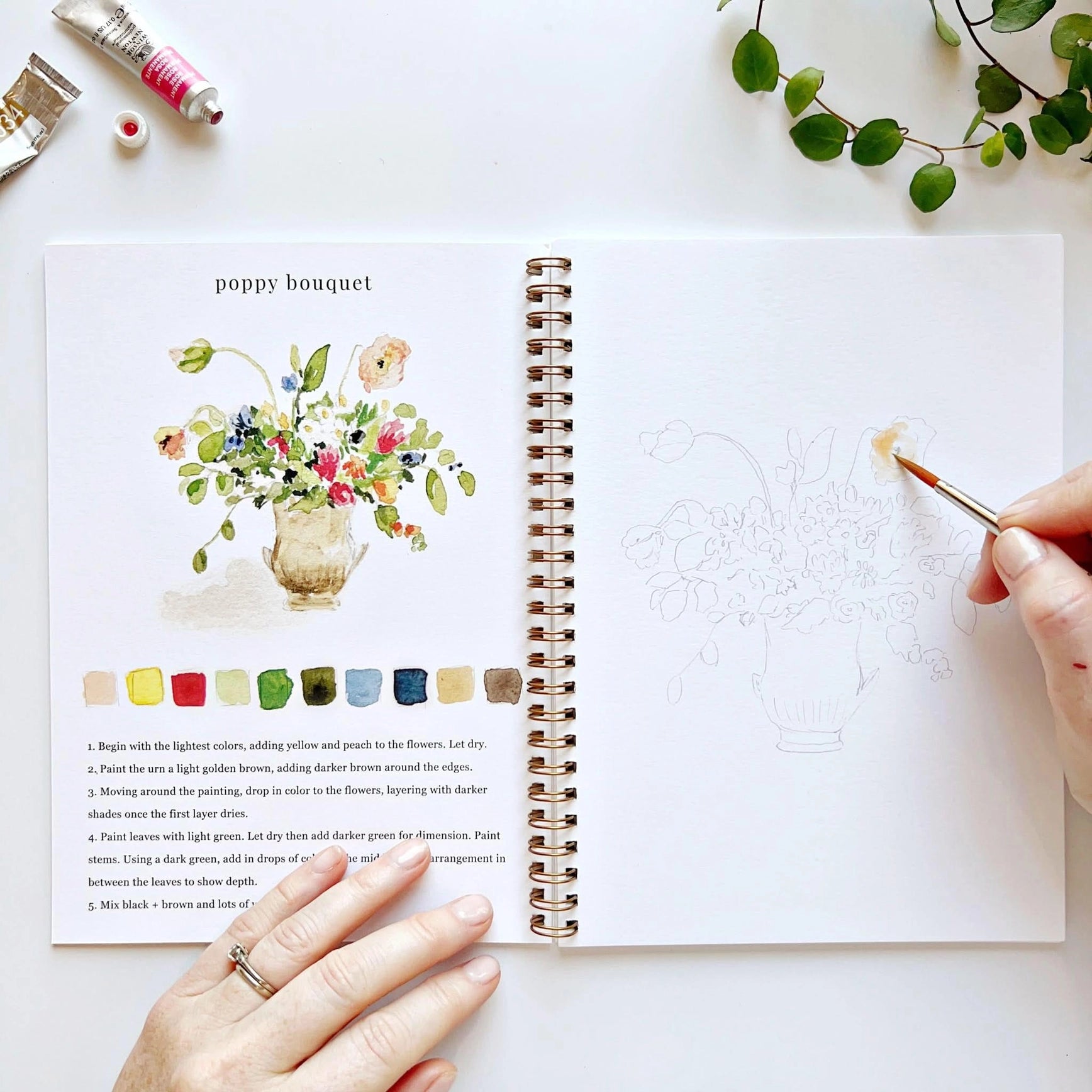 Open book with a painting of a poppy bouquet and a hand holding a paintbrush.