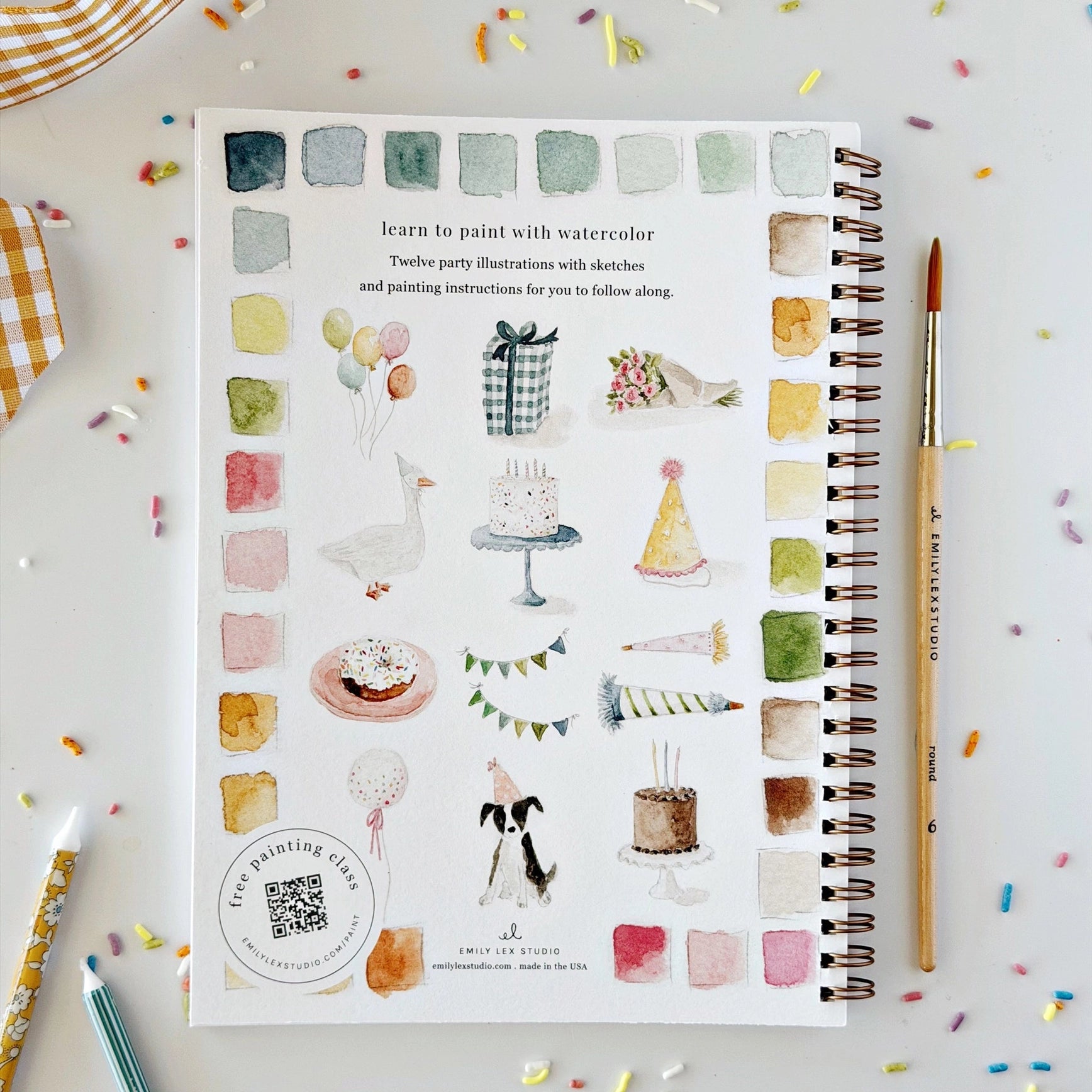 A spiral-bound watercolor painting guide book titled 'learn to paint with watercolor' is displayed on a white surface with colorful confetti. The book cover features various watercolor illustrations of birthday-themed subjects like cakes, balloons, and party hats, alongside a column of watercolor swatches.