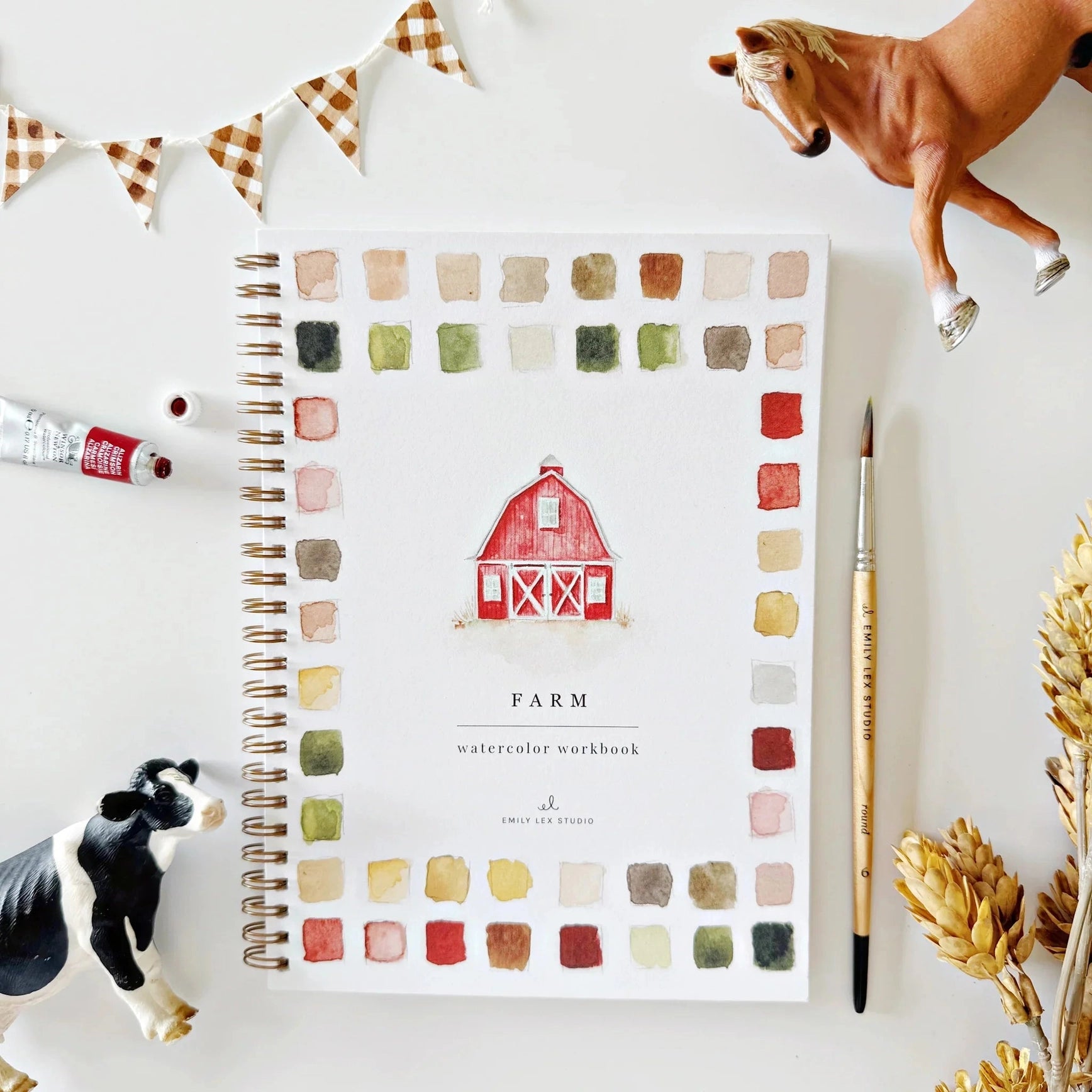 A spiral-bound watercolor workbook titled 'FARM' lies on a white surface, surrounded by farm-themed props including a toy horse, cow, paintbrush, paint tube, wheat ears, and a bunting garland. The cover shows a red barn illustration framed by watercolor swatches in muted earthy tones.