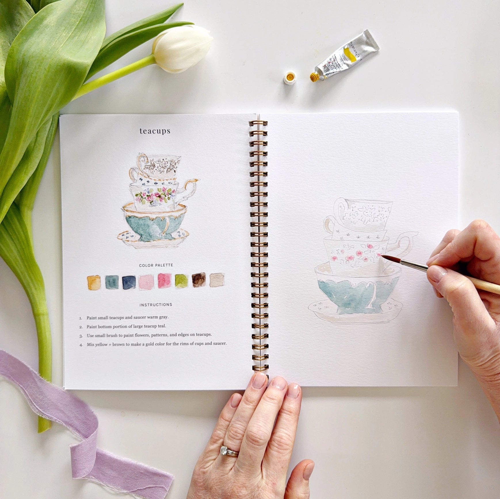 Person drawing teacups in a sketchbook with a tulip and paint tubes on a white surface