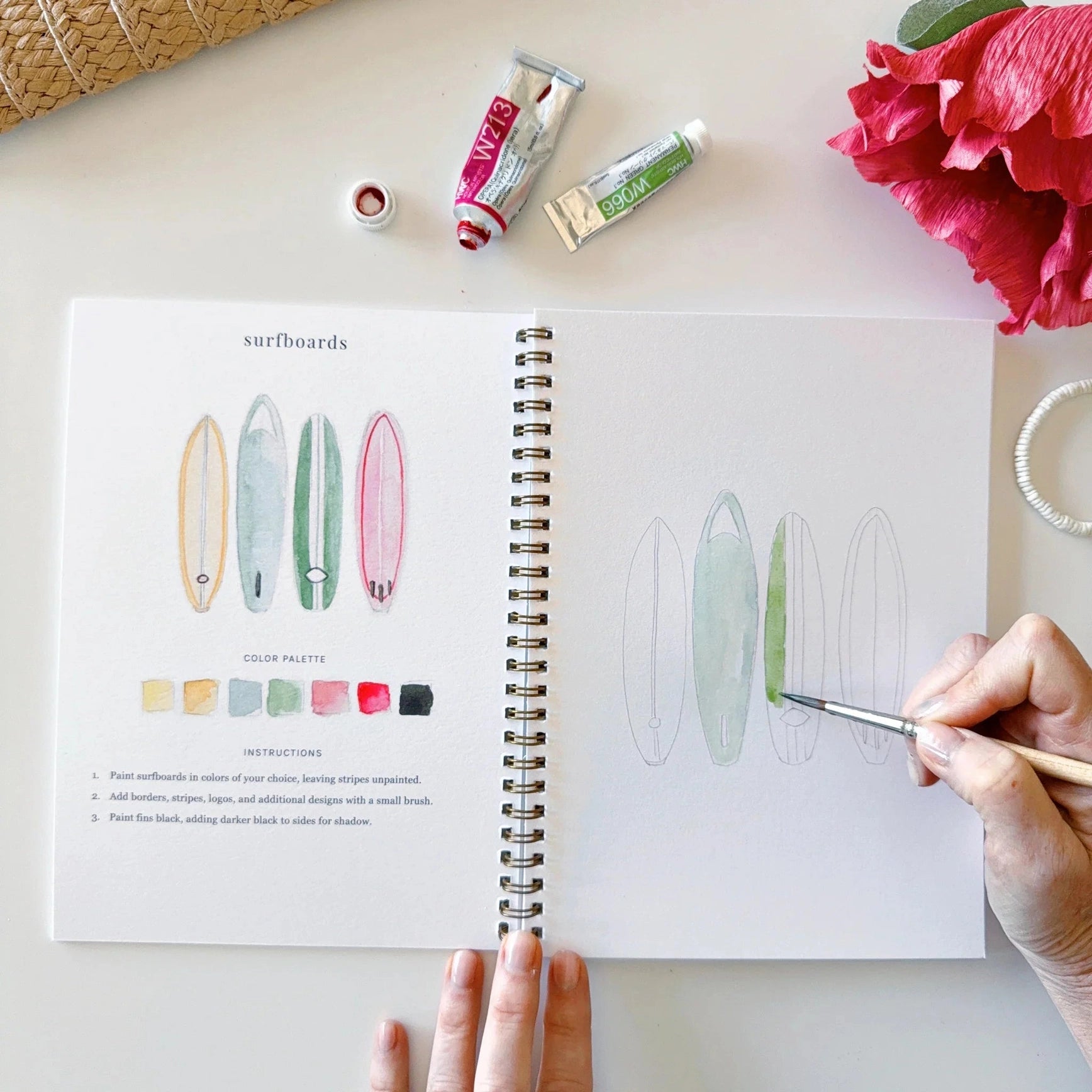 Person painting a design in a sketchbook with art supplies on a table.