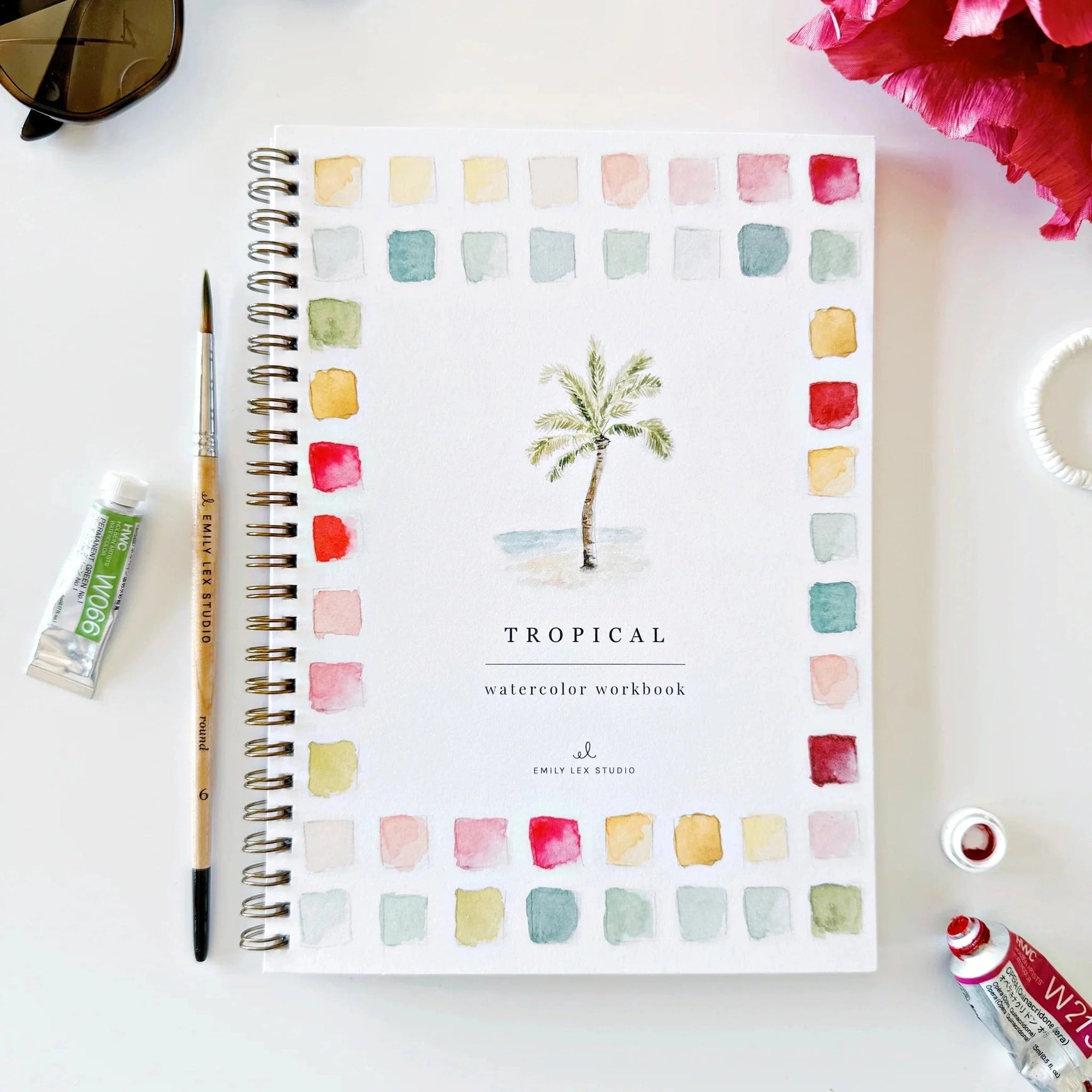 A spiral-bound watercolor workbook titled 'TROPICAL watercolor workbook' by Emily Lex Studio, displayed on a white surface with art supplies including a paintbrush, paint tubes, sunglasses, and flowers.