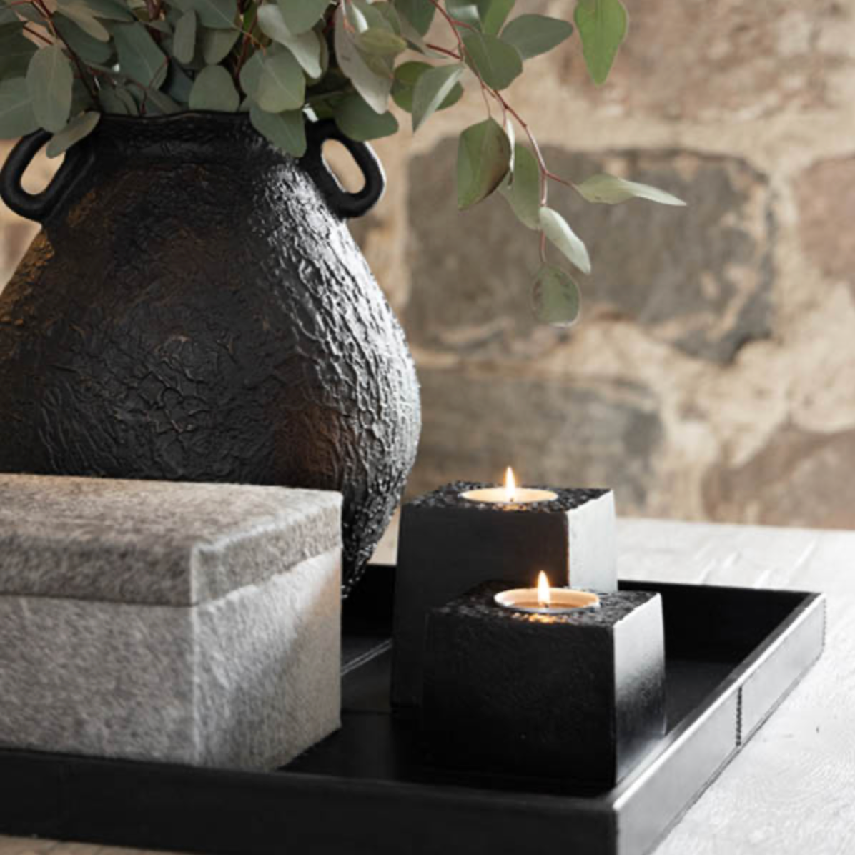 Decorative black tray holding two lit black cube candle holders with pebbles, a gray textured box, and a large black textured vase with eucalyptus branches, set against a rustic stone wall background.