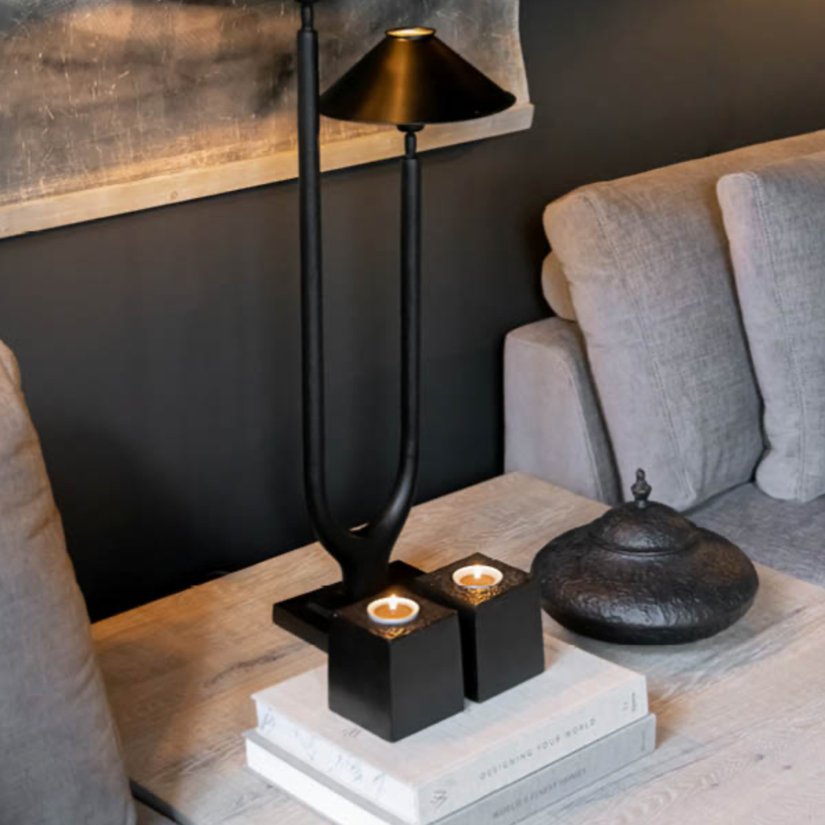 A modern black table lamp with a dual-branch design and conical shades, styled on a light wood table next to a gray sofa. The lamp is displayed on stacked books alongside two black candle holders with lit candles and a dark textured decorative pot.