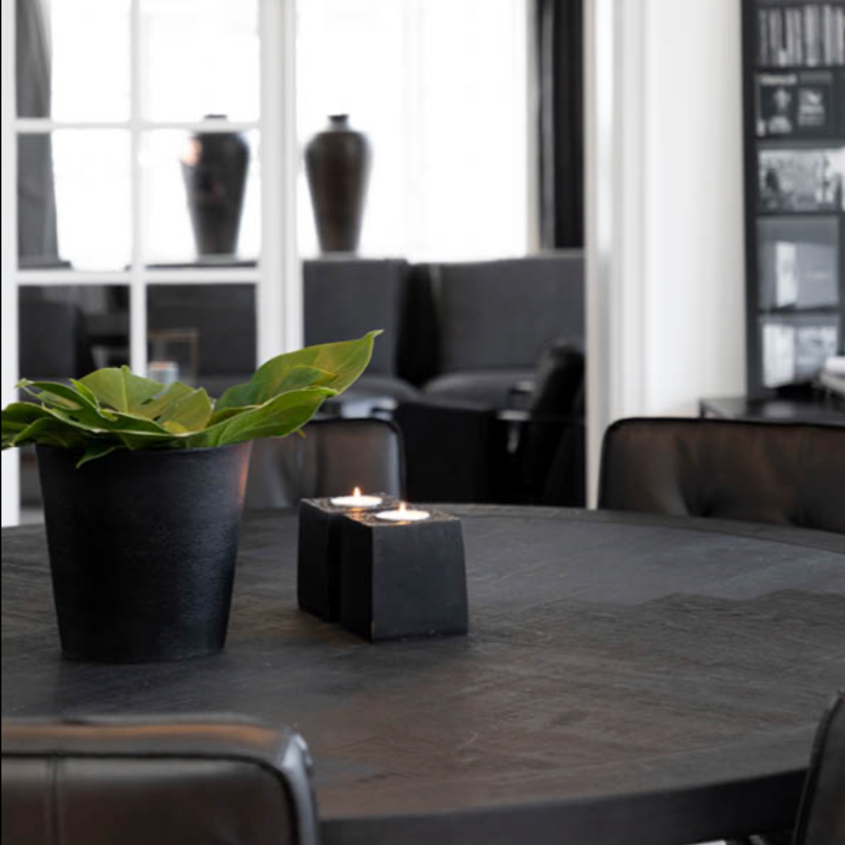 A modern black ceramic candle holder with two lit tealights sits on a dark round table next to a black planter with green leaves in a stylishly decorated interior room.