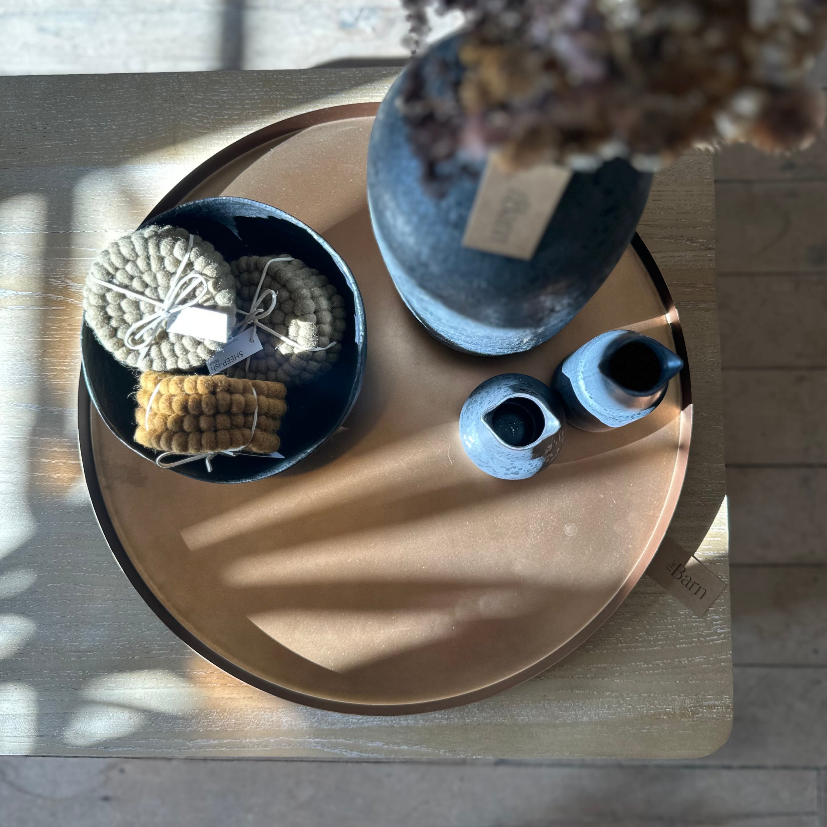 An overhead styled shot of a rustic ceramic table setting on a light wood floor. The centerpiece is a large round copper-toned tray holding a dark blue bowl with knitted beige and yellow corn-cob-shaped items, a matching blue vase with dried flowers, and two small blue ceramic vessels.