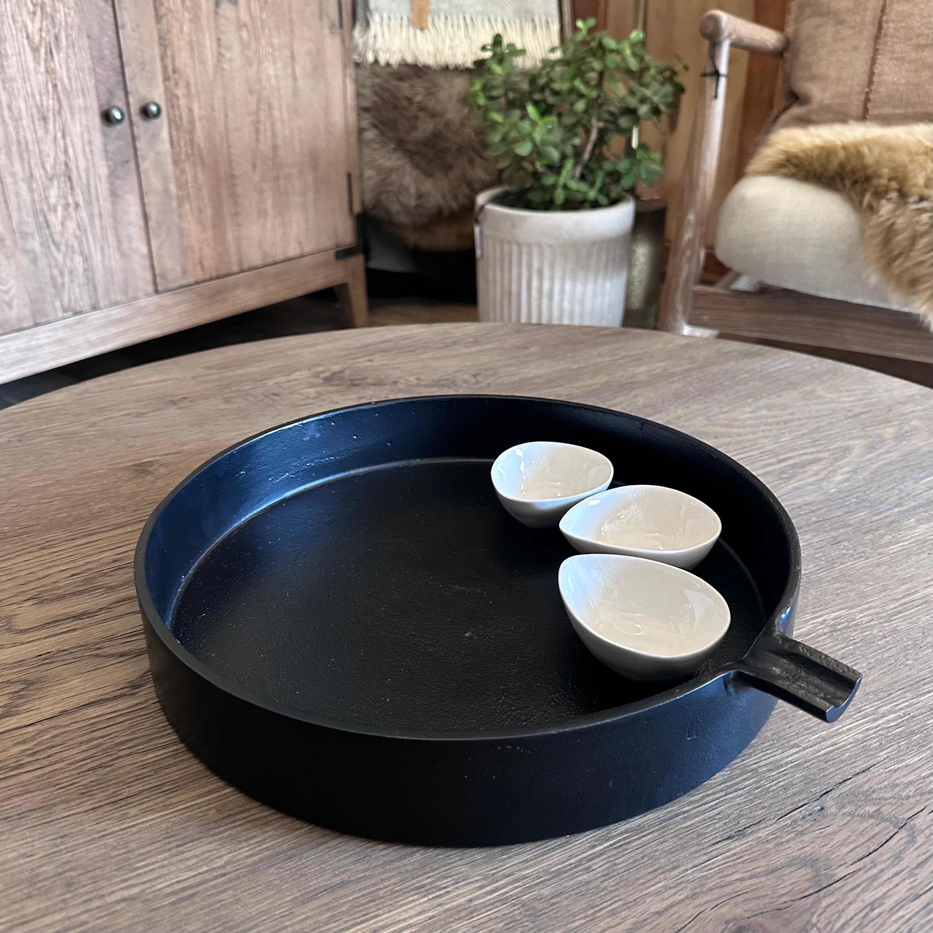 A round black metal tray with a small handle, displayed on a wooden table. Inside the tray are three small white ceramic cups.