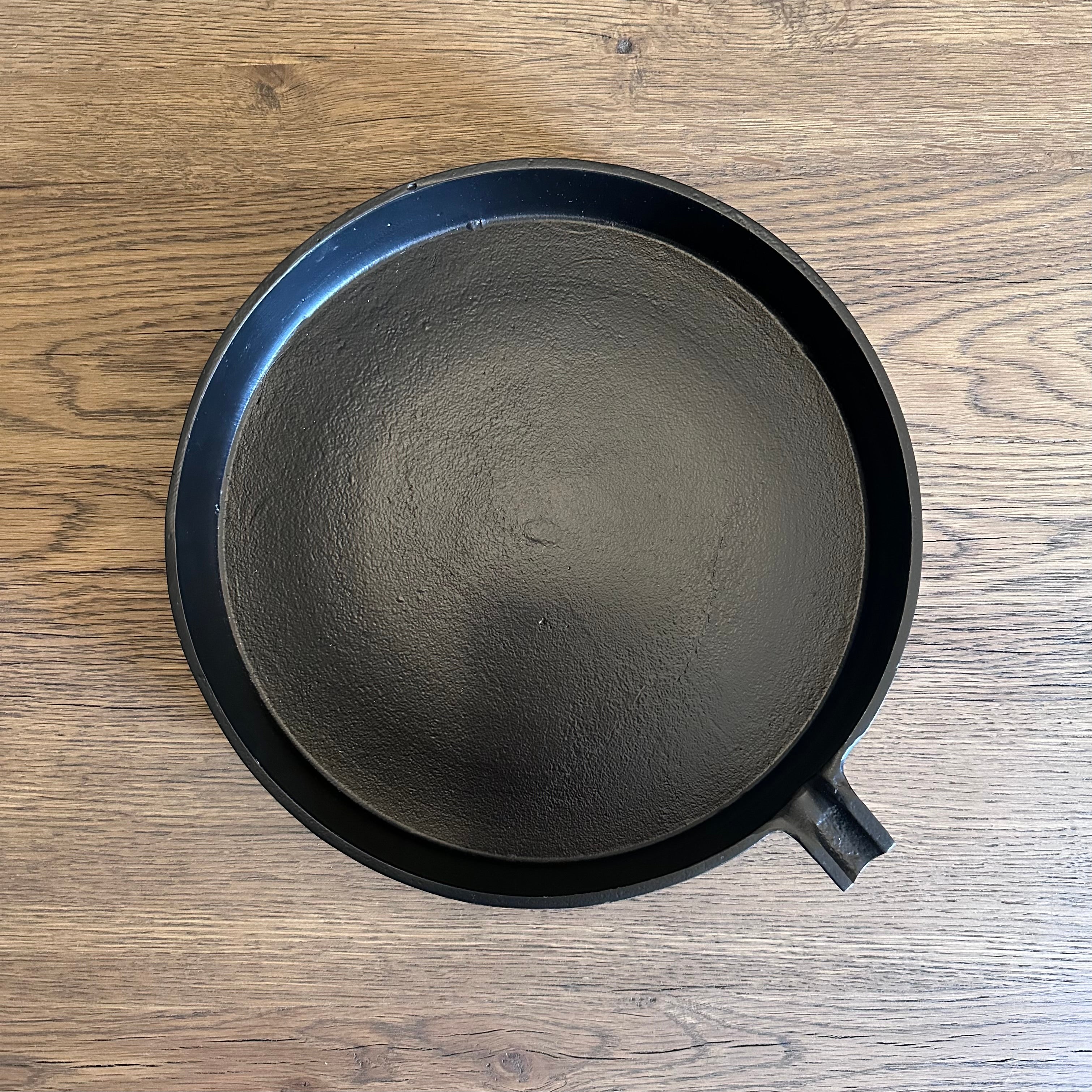 A top-down view of a black cast iron skillet with a small handle, resting on a light brown wooden surface.