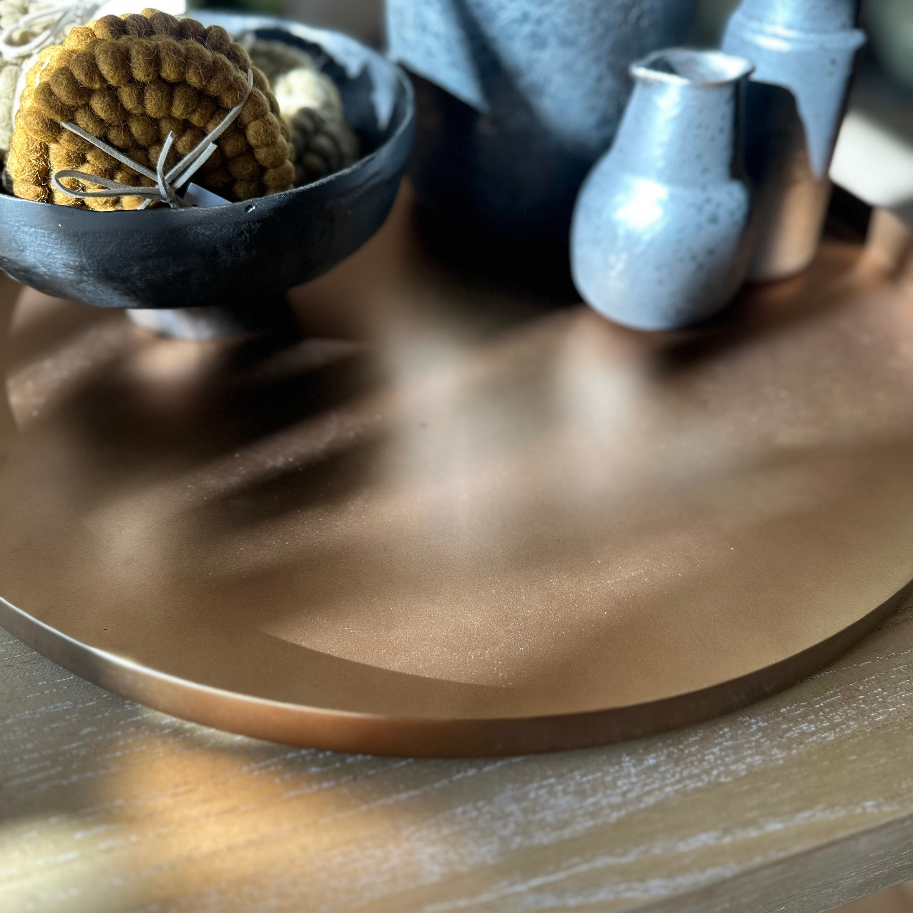 A close-up of a round, brushed copper or bronze tray resting on a wooden surface, with a dark bowl and small ceramic vessels in the softly blurred background.