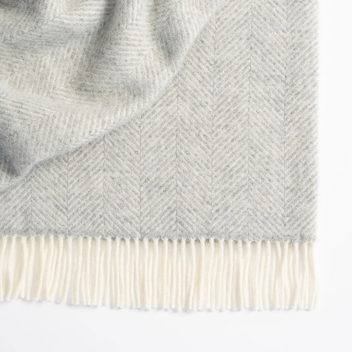 A close-up of a light gray and off-white herringbone patterned throw blanket with cream-colored fringe at the bottom, shown against a plain white background.