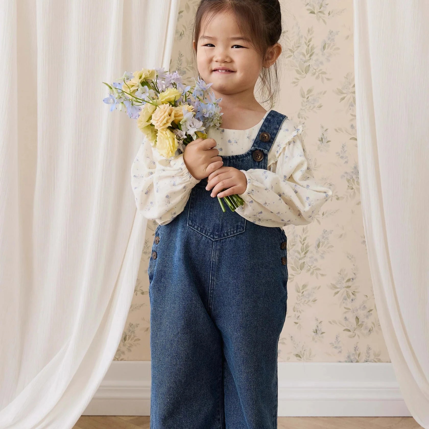 A young child wearing a pair of wide-leg blue denim overalls over a cream lace-up blouse, holding a small bouquet of yellow and blue flowers. The child is standing in a room with light wood floors, cream drapes, and floral wallpaper.