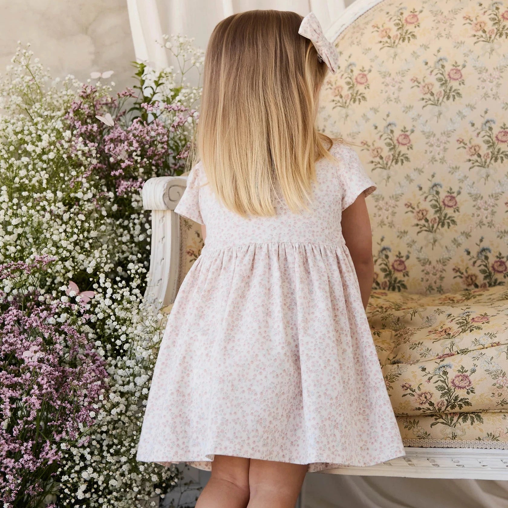 Young girl in a floral dress standing next to a floral arrangement in a room with vintage furniture.