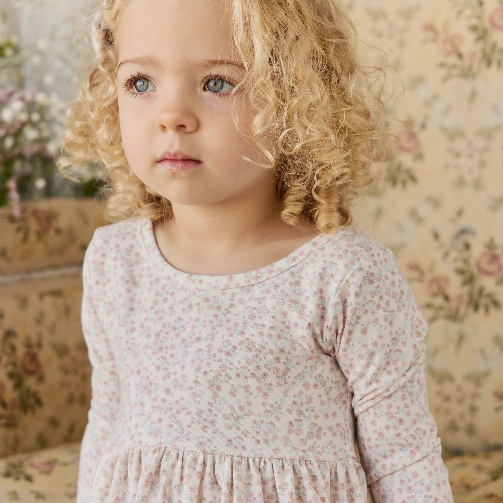 Young girl in a floral dress standing in a room with floral wallpaper.