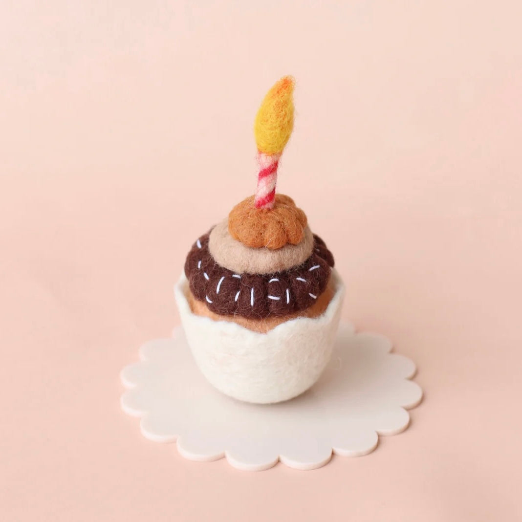 Felted toy resembling a birthday cupcake with a candle on a white plate against a beige background