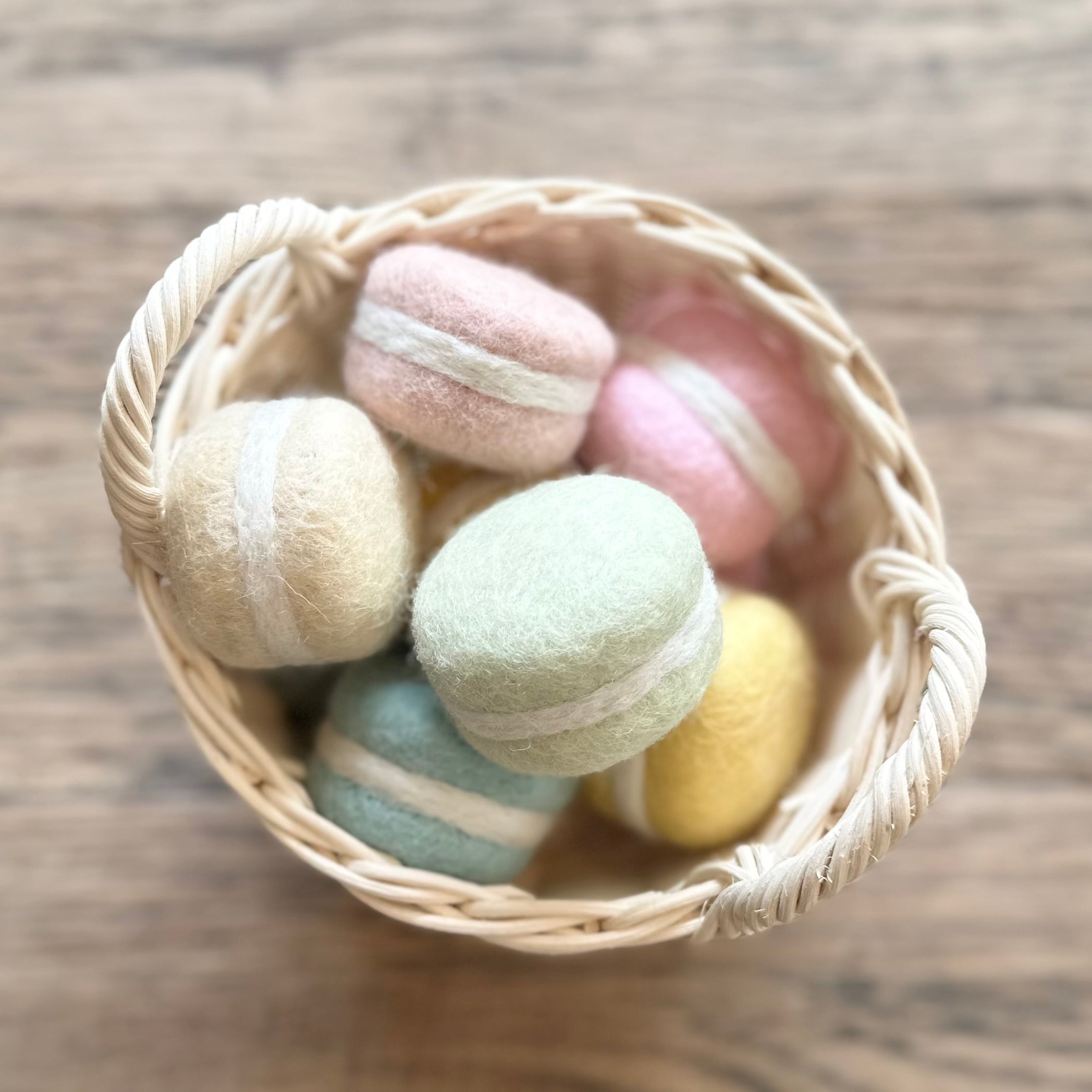 A small woven basket filled with pastel macaron-style felt items in cream, pink, mint green, blue, and yellow, photographed on a wooden surface.