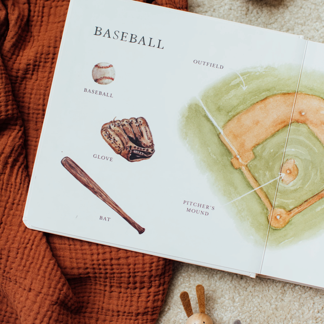 An open children's book spread showing baseball-themed illustrations, including a baseball, glove, bat, and a watercolor field diagram, styled on a rust-colored textured blanket with a beige carpet visible around it.