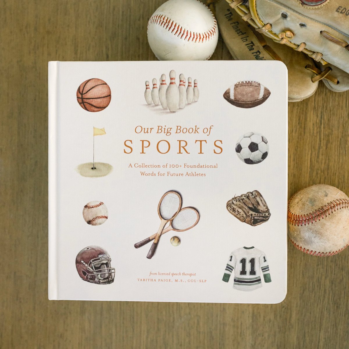 Our Big Book of Sports children's book displayed on a wooden surface surrounded by baseballs, a glove, and a helmet.