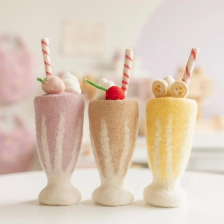 Three miniature felt milkshakes in pastel pink, beige, and yellow, each topped with whipped cream, a cherry, a candy corn, and a striped paper straw, arranged on a white surface against a soft-focus pastel background.