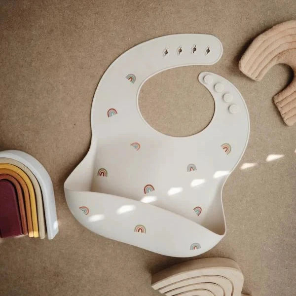 A white silicone baby bib with small rainbow prints, laid flat on a textured brown surface with wooden toy arches nearby.
