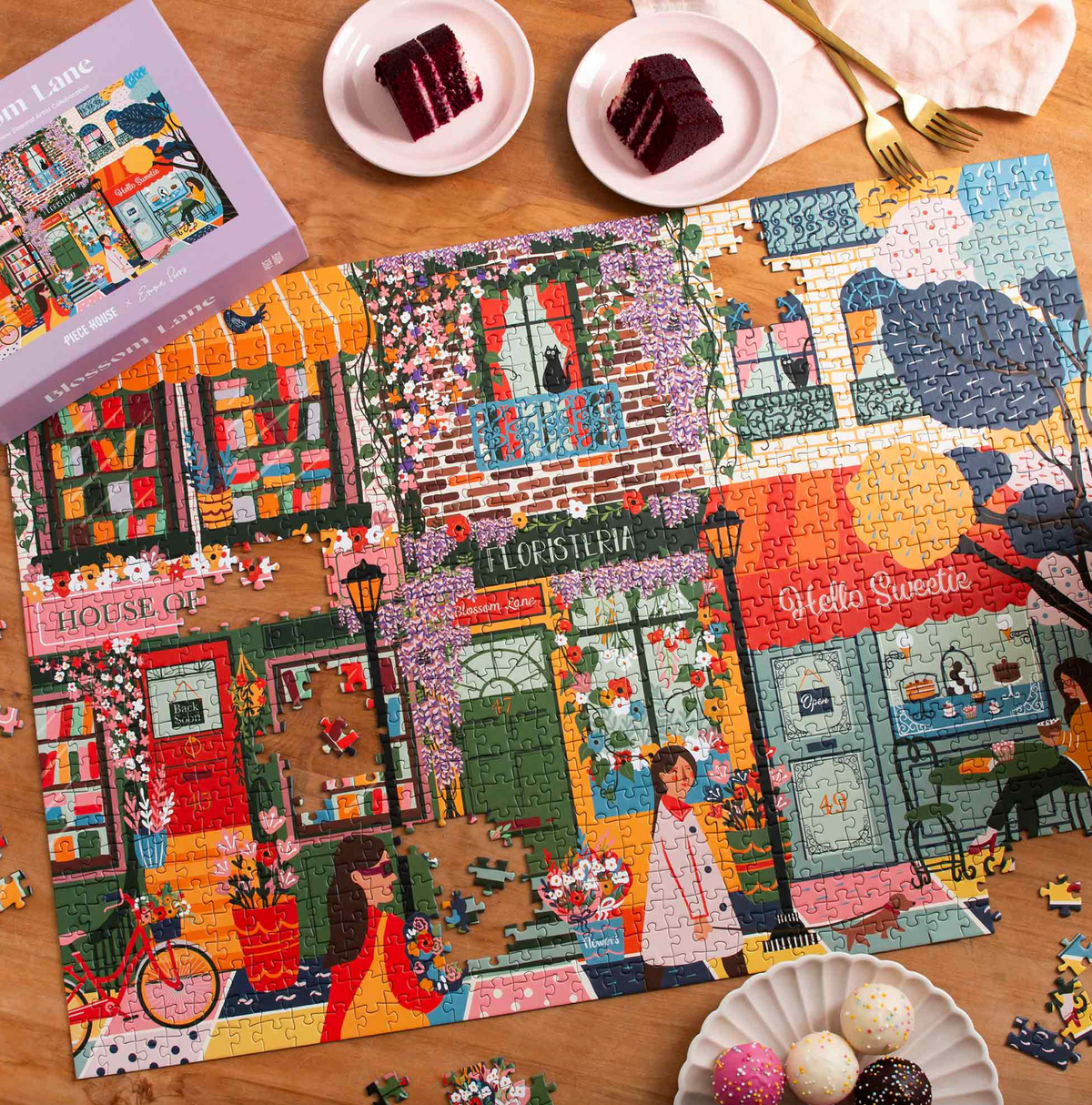 A completed multicolor jigsaw puzzle showing a whimsical street scene with shopfronts, flowers, bicycles, and characters, arranged on a wooden table. The partially cut puzzle pieces are scattered around it, along with a purple puzzle box, small plates with cake slices, and decorative chocolates.