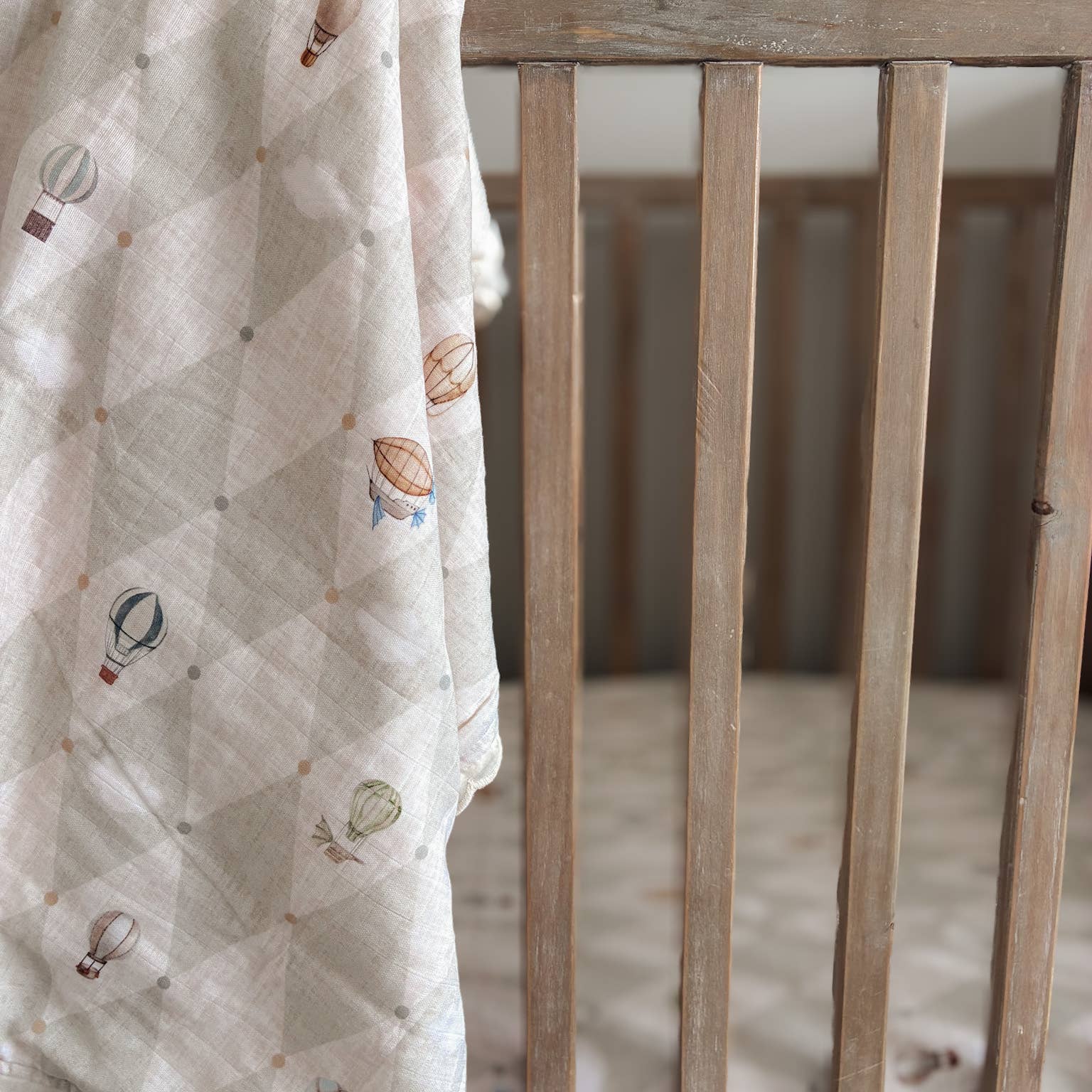 A close-up of a baby blanket draped over the slats of a wooden crib. The blanket is a light cream color with a diamond grid pattern and small illustrations of hot air balloons in muted earthy tones.