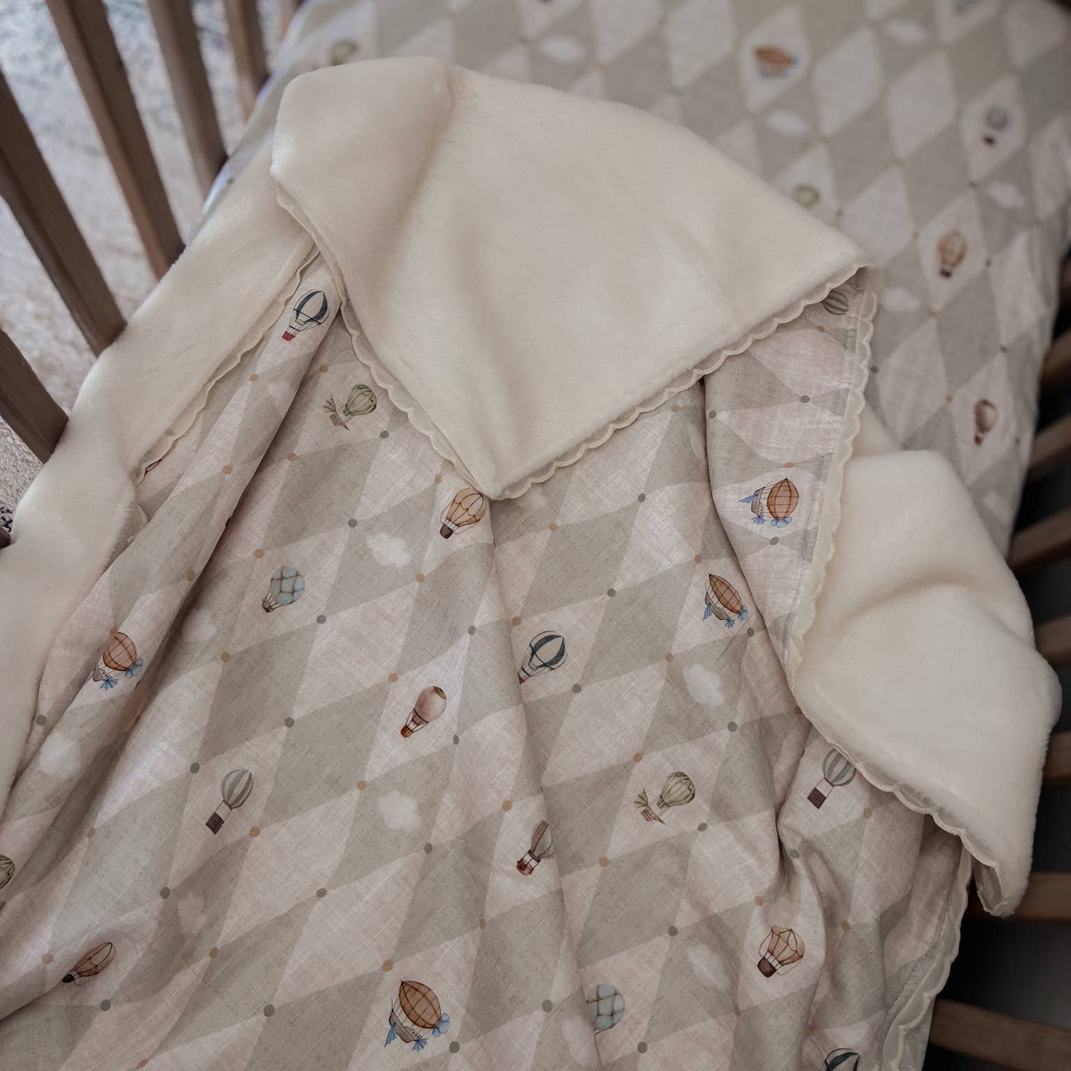 A close-up of a baby blanket draped over a crib, featuring a light beige and gray argyle pattern with small hot air balloon prints and a soft cream fleece lining.