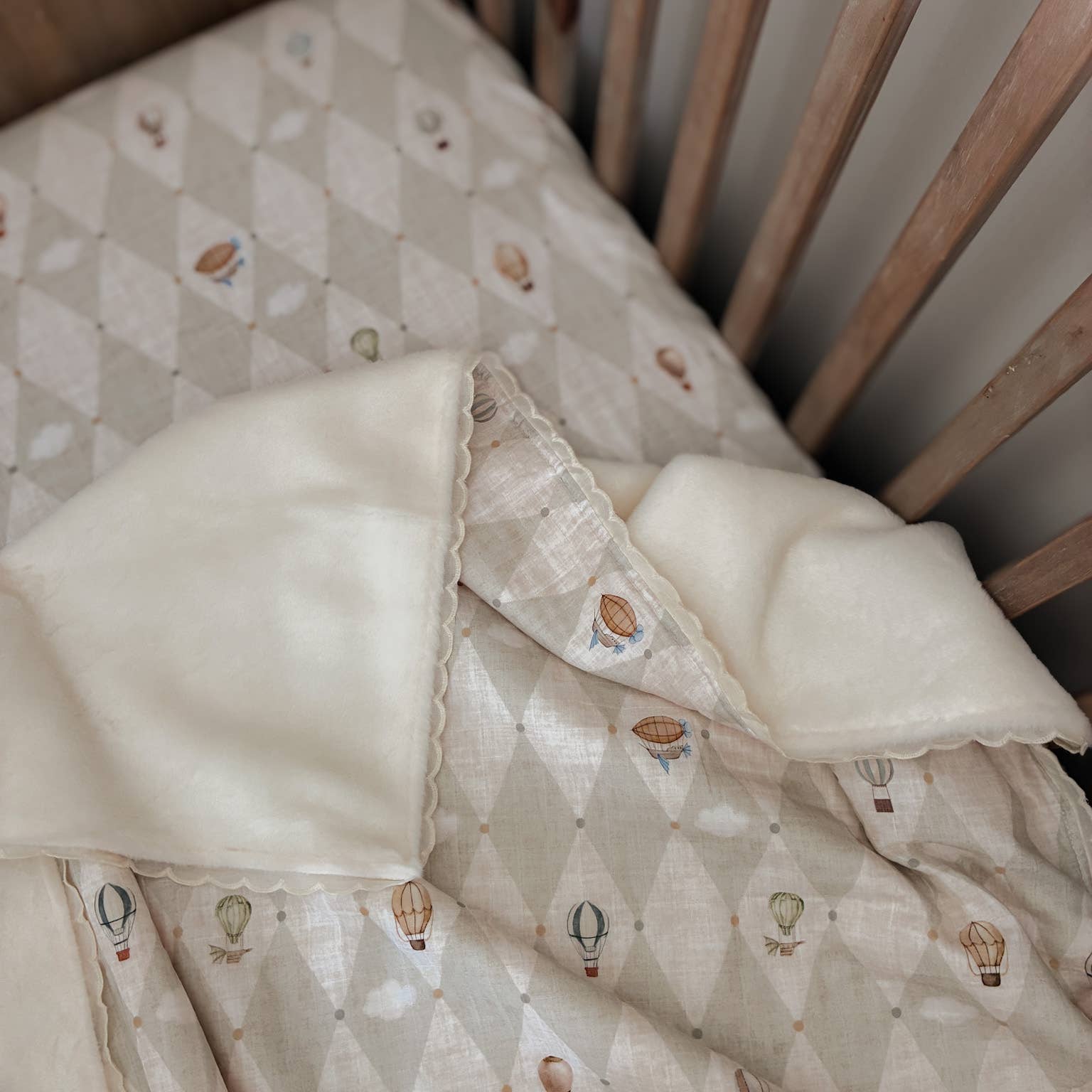 A close-up of a baby bedding set in a wooden crib, featuring a cream-colored plush blanket with scalloped edges layered over a diamond-patterned quilt with a hot air balloon print.