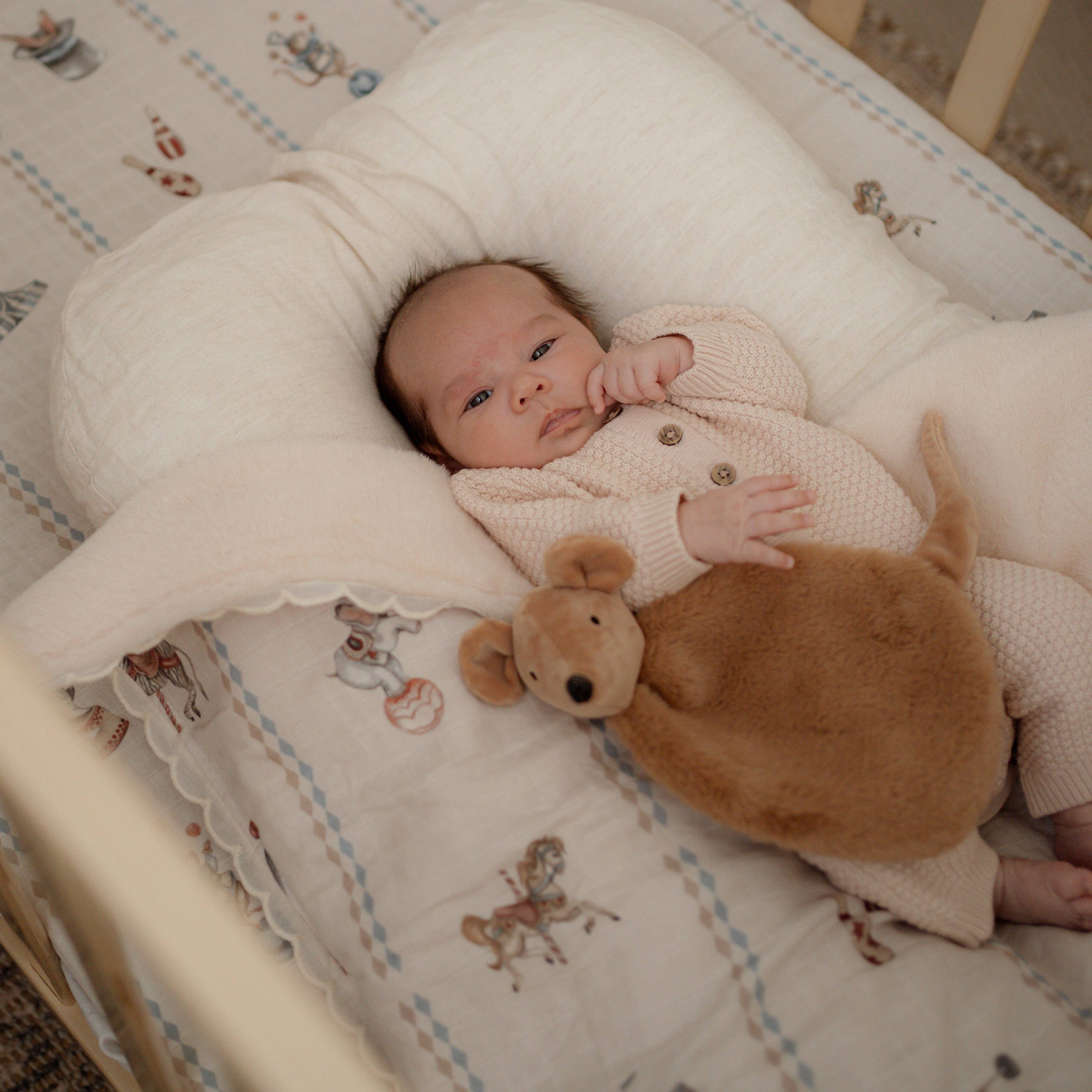 A baby lying on a cream-colored nursing pillow in a crib, wearing a light pink knit romper and holding a brown plush kangaroo toy.