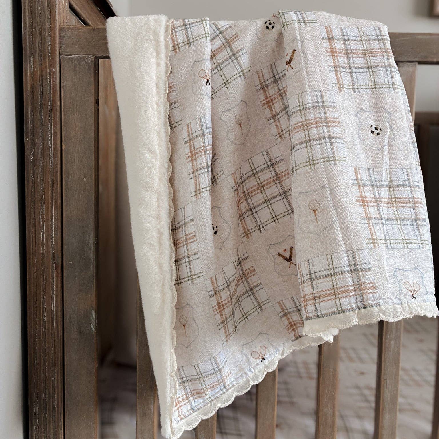 A soft baby blanket draped over a wooden crib, featuring a cream-colored plush fleece border and a patchwork-style printed cotton front with plaid squares, embroidered bunny motifs, and small bow details.