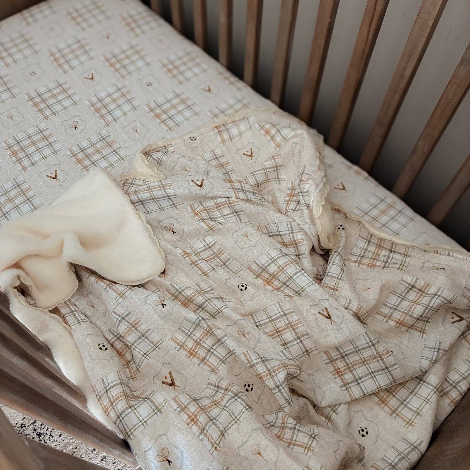 A baby bedding set including a plaid muslin blanket and a soft cream-colored swaddle, styled inside a wooden crib.