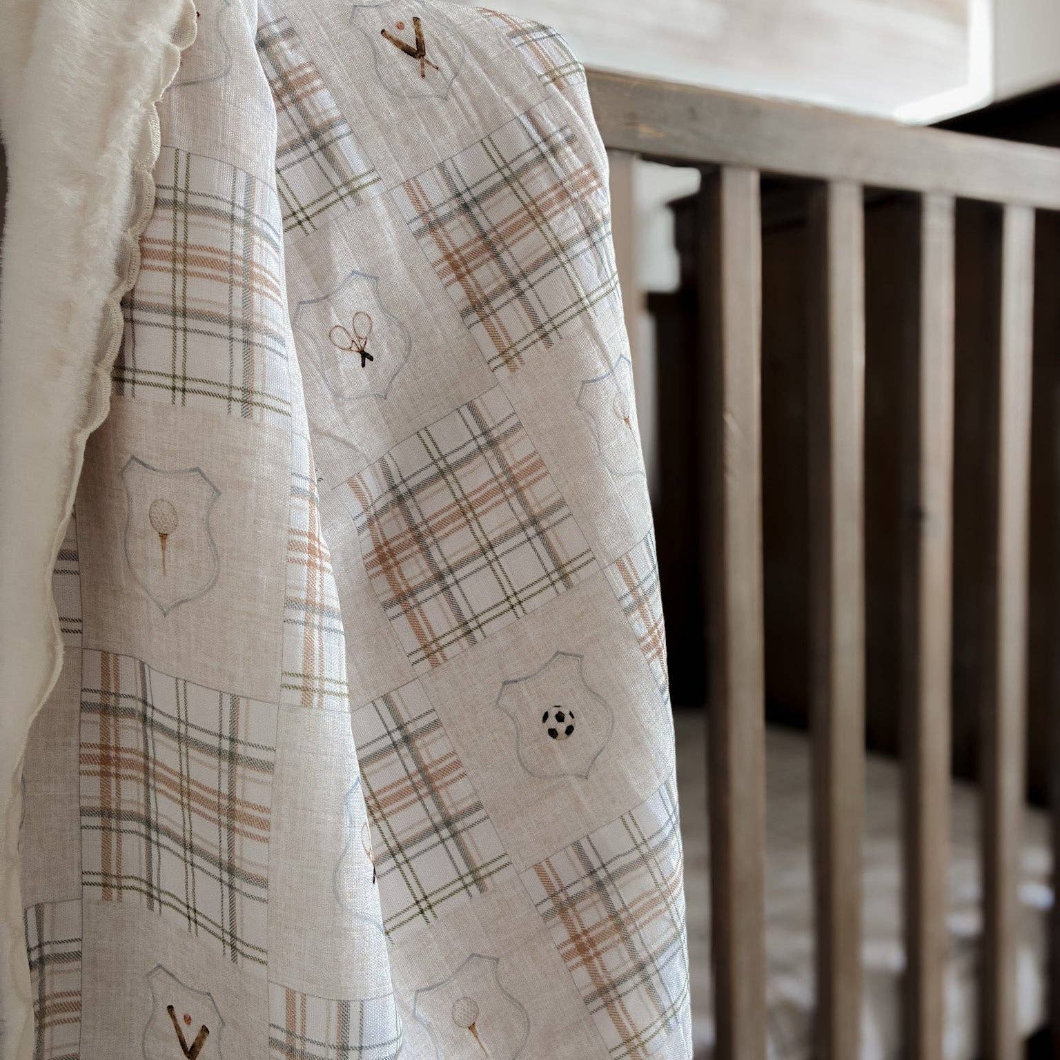 A close-up of a soft muslin baby blanket draped over a wooden crib. The blanket has a warm cream base with a plaid pattern in muted brown and gray, decorated with embroidered shield motifs and small sports-themed symbols.