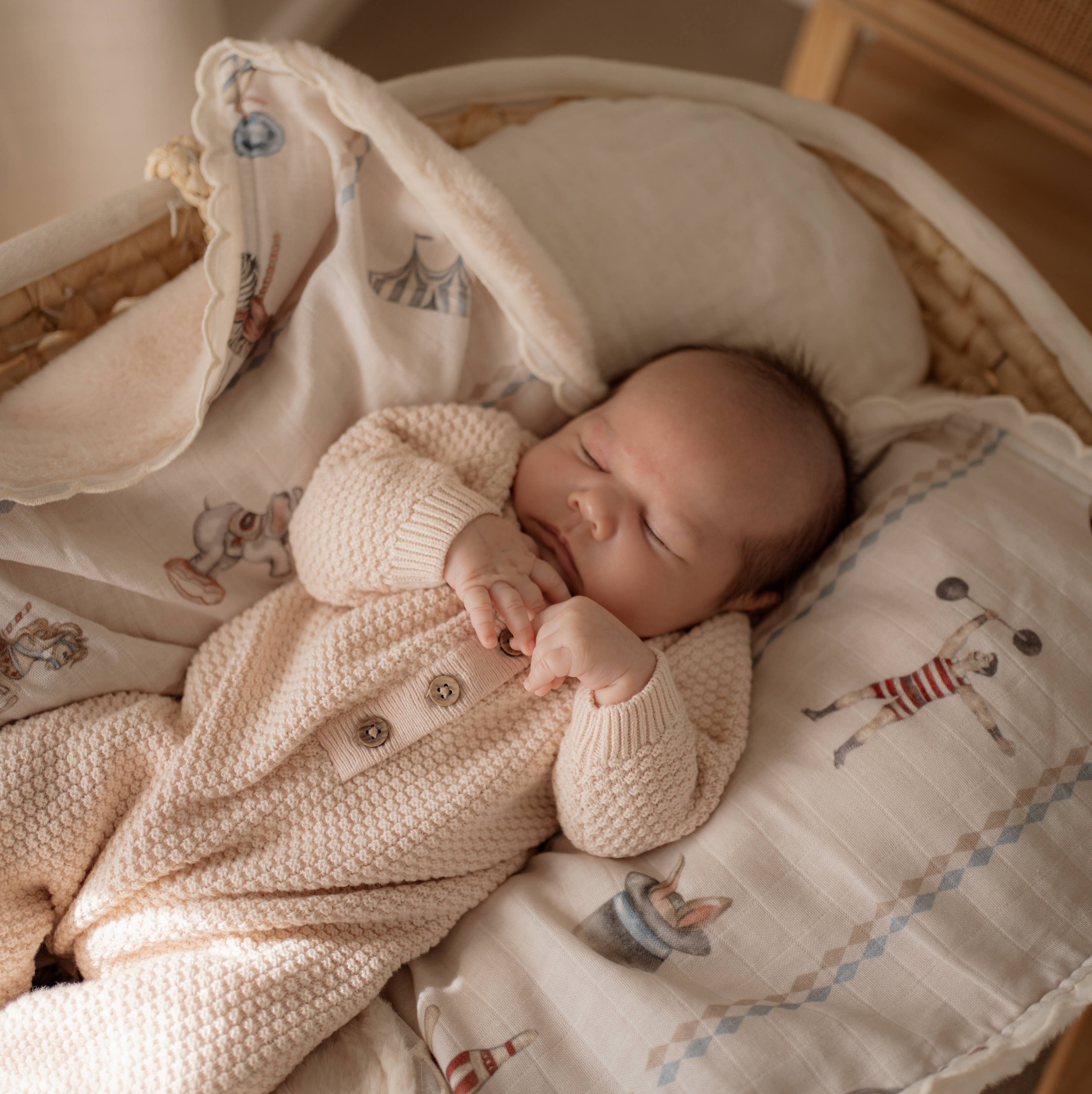 A sleeping newborn baby wearing a cream-colored textured knit romper, lying in a wicker bassinet with matching patterned muslin blankets and pillows.